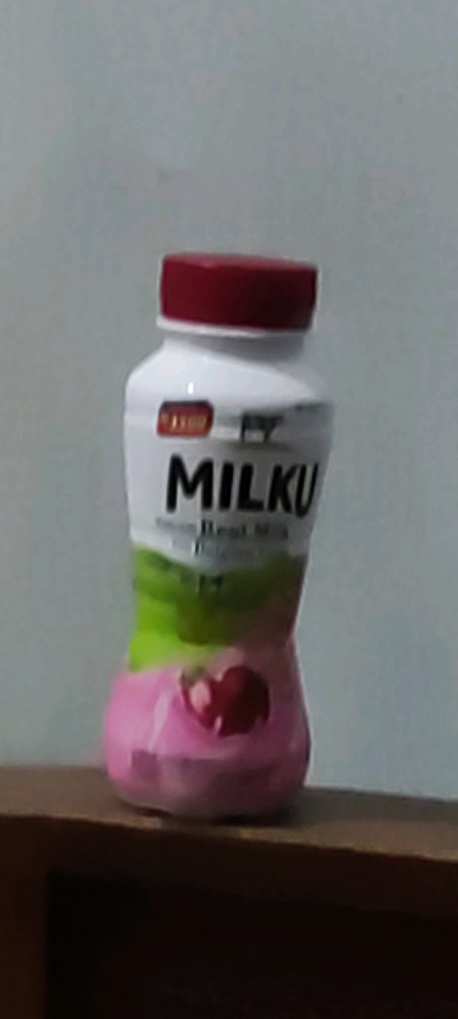 A white plastic bottle with a red cap is placed on a wooden surface. The bottle has black text that reads "MILKU". Below "MILKU," there is smaller text that is difficult to read due to low resolution but appears to say "Real Milk". The bottle has a gradient design with green and pink colors. A red cherry graphic is visible on the pink portion of the bottle. The background is a plain, light-colored wall. The image is blurred and grainy, indicating it was taken with a low-quality camera or in poor lighting conditions. There are no people or specific environmental features that would indicate a location within the city of Karangwangun, Indonesia.