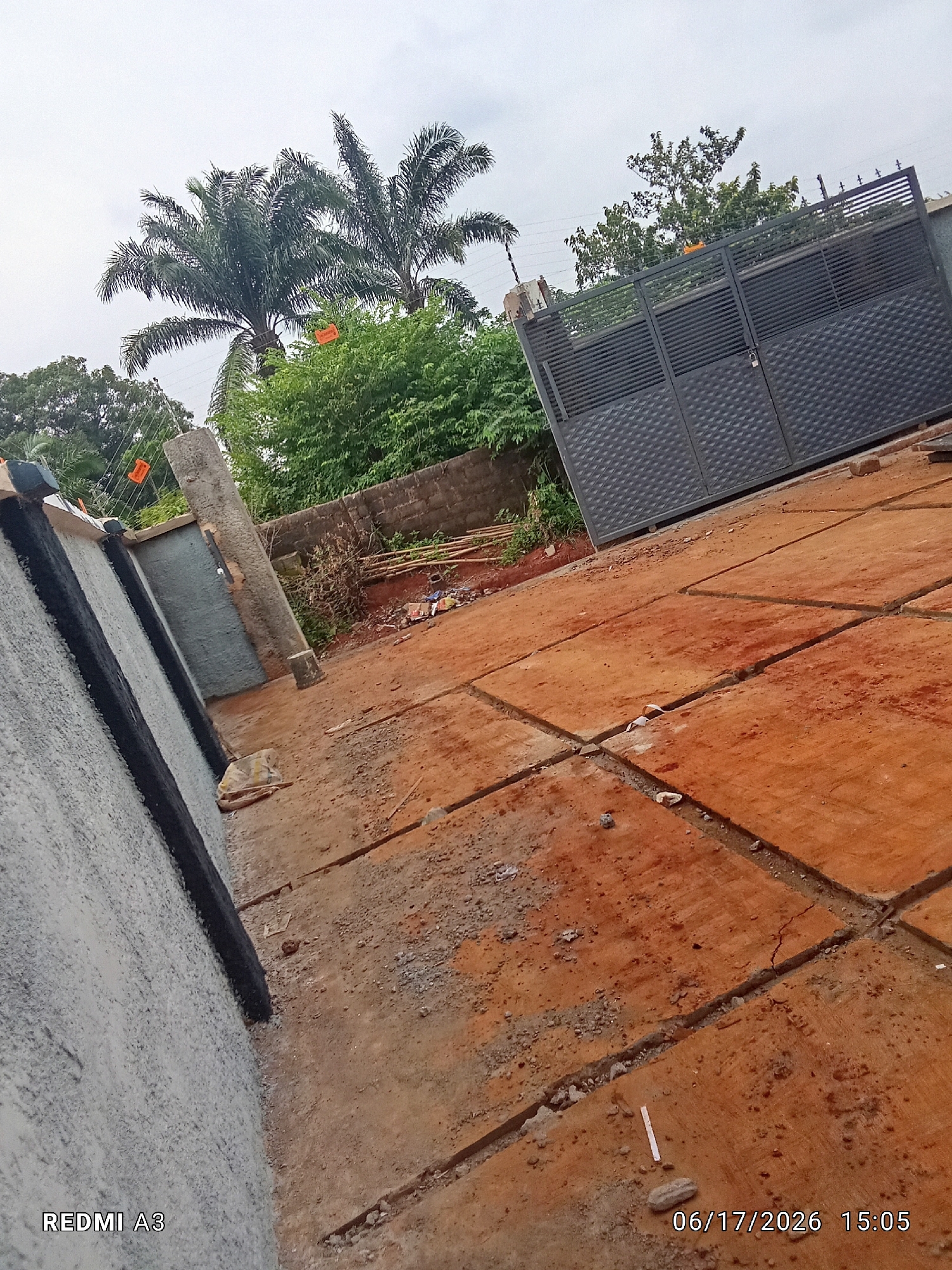 The image depicts an outdoor scene at what appears to be a construction site or a newly paved area in Nawfia, Nigeria. The primary subjects are a large, dark grey metal gate with a diamond pattern and an attached lock, positioned on the right side of the frame. In the foreground and extending to the left, a grey wall with black stripes is visible. The ground is covered with reddish-brown soil and appears to be paved with square slabs, some of which show cracks and are separated by trenches.
Behind the wall, a concrete pillar stands, and further back, dense green foliage and palm trees are visible. The sky is overcast, suggesting a cloudy or possibly rainy day, and the lighting indicates it is daytime, likely late afternoon given the timestamp. There are no people visible in the image.
Notable details include the unfinished nature of the ground paving, with exposed earth and trenches. The presence of the electric fence wires on top of the gate and wall, along with orange warning signs, suggests a security measure. The overall impression is one of ongoing construction or development.
Visible text includes "REDMI A3" in the bottom left corner and a timestamp "06/17/2026 15:05" in the bottom right corner.
