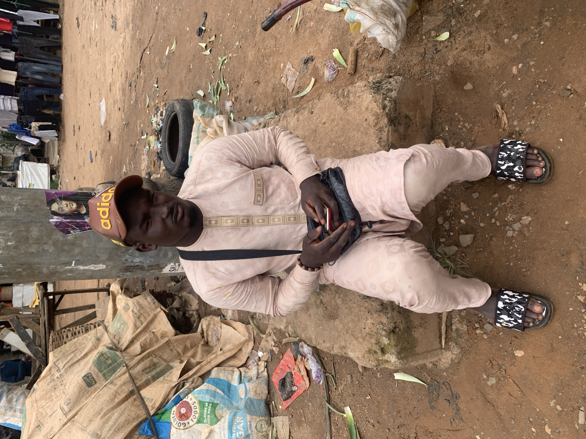 The image depicts a young Black man sitting on a concrete block in an outdoor market setting, likely in Abuja, Nigeria, given the context. He is wearing a light pink traditional African garment, possibly a kaftan, with intricate embroidery around the neckline and down the front. On his head, he has a brown baseball cap with yellow lettering that appears to say "adidas". He is looking towards the camera with a neutral expression and has a black crossbody bag slung over his shoulder, with his hands inside the bag, holding a red smartphone. He is wearing black and white patterned slides on his feet.
The environment around him is somewhat cluttered and appears to be a bustling marketplace. To his left, there are large, possibly burlap sacks, one with a blue and white label that reads "GOLDEN FOODS". A discarded red package and other debris are scattered on the ground. Behind him, there is a large concrete pillar with posters, one of which features a woman's face. Further in the background, tires, more sacks, and a display of clothing hanging on a rack are visible. The ground is covered in red earth, with scattered leaves and debris. The overall impression is of a vibrant, working market.