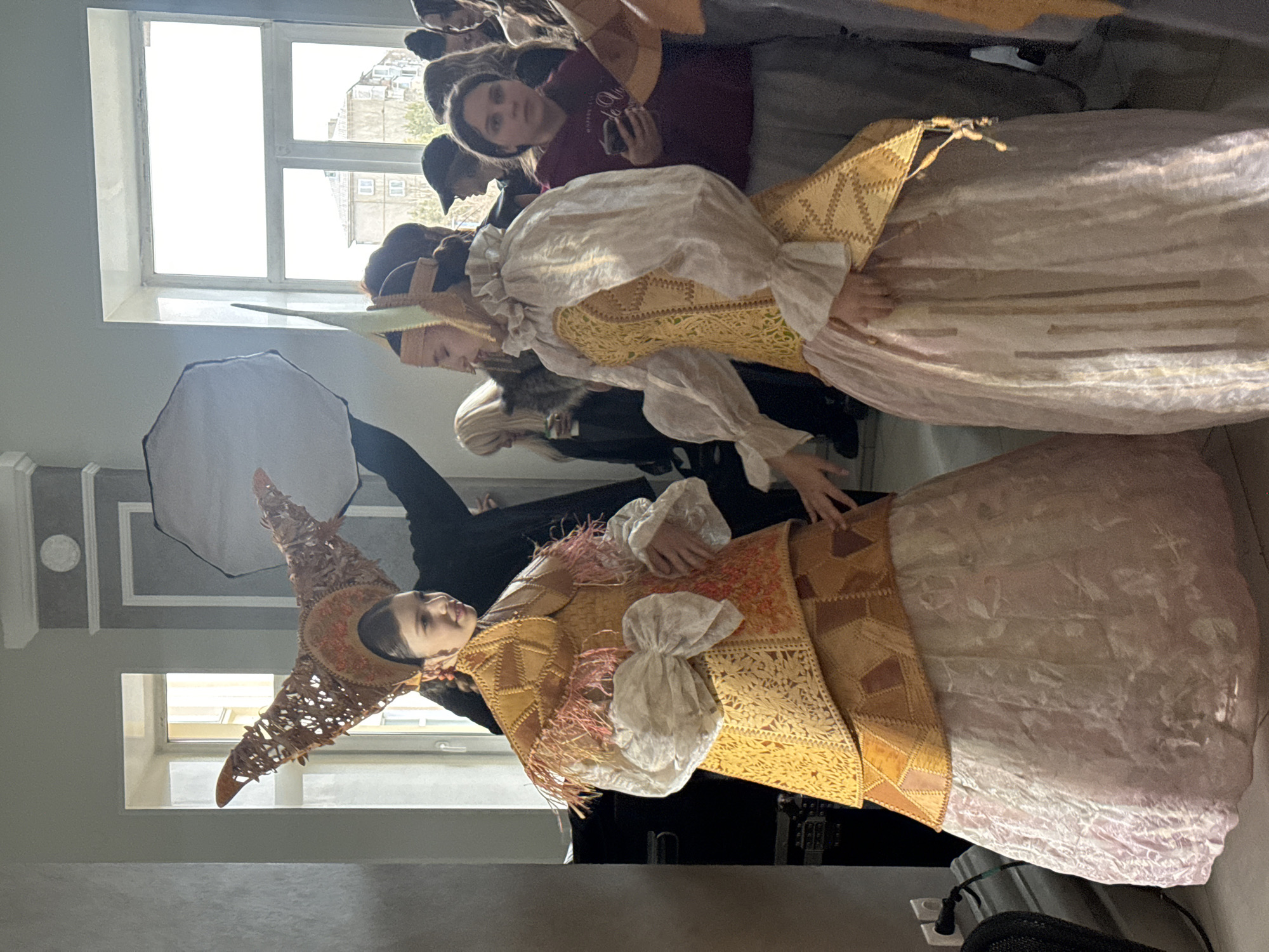 The image shows two women dressed in elaborate, period-style costumes. One woman wears a tall, conical hat and a yellow and orange patterned dress with a large white bow. The other woman wears a simpler headpiece and a yellow and white dress. A group of people is gathered behind them, seemingly observing or participating in a photoshoot. The setting appears to be indoors, near a window, with lighting equipment visible. The location is Unknown, Unknown.
