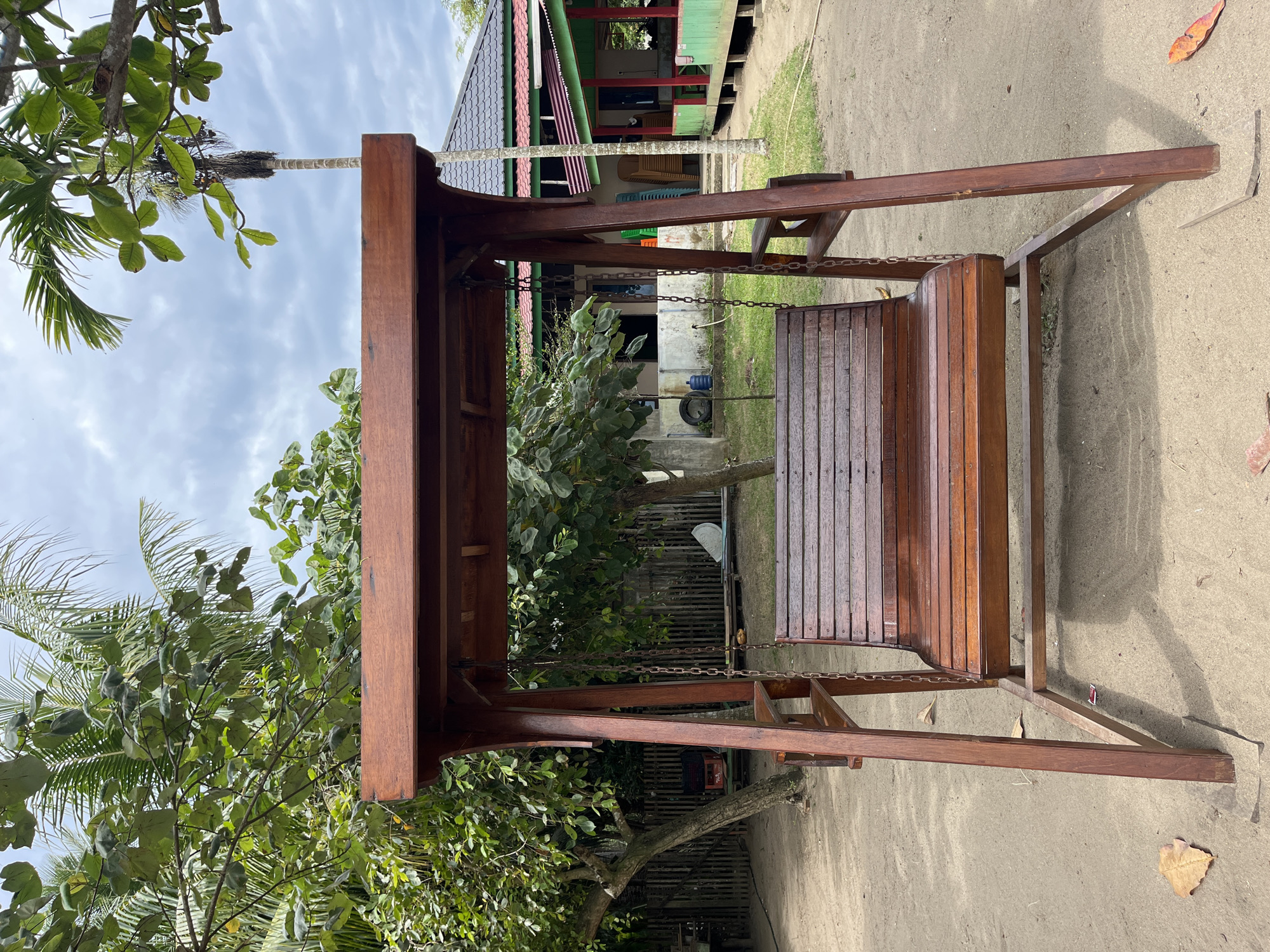 The image depicts a wooden swing set outdoors, likely in a park or residential area of Lhokseumawe, Indonesia. The swing is made of stained wood with a seat suspended by chains. The ground is sandy or dirt. The background includes a house with green and red trim. Vegetation, including trees and foliage, is visible on both sides of the swing. The sky is cloudy. No people are visible in the frame.