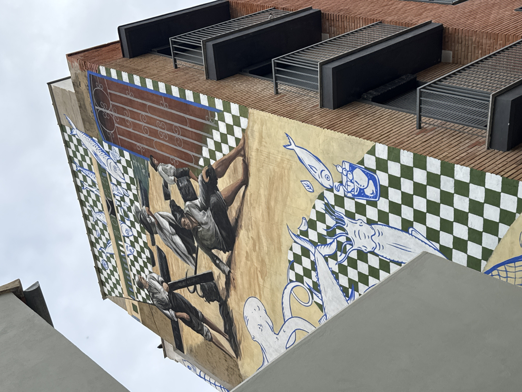 An angled, upward-looking shot captures the corner of a building in València, Spain, adorned with a vibrant and complex mural. The mural spans a significant portion of the building's side, featuring human figures in a dynamic, classical style. One figure, centrally positioned, appears to be reaching or falling, while another lower down seems to be interacting with a dark, cross-like structure. Their expressions suggest exertion or drama.
The artwork incorporates various patterns and marine life. Sections of the mural feature green and white checkerboard designs, and stylized blue and white depictions of fish, including mackerel, an octopus, and other aquatic creatures, some of which are placed against a light brown, parchment-like background. An upper section of the mural displays a deep reddish-brown panel with white, swirling linear motifs that resemble musical staves or abstract patterns.
Above the mural, the building transitions to a facade of reddish-brown bricks arranged in neat horizontal lines. Several modern, dark-colored rectangular structures with slatted grates are affixed to the upper part of the building, possibly serving as architectural vents or protective coverings for windows. Parts of a plain light gray building or wall are visible in the foreground and bottom-right, framing the main subject. The sky is overcast, indicating a daytime scene. No discernible text is visible within the image.