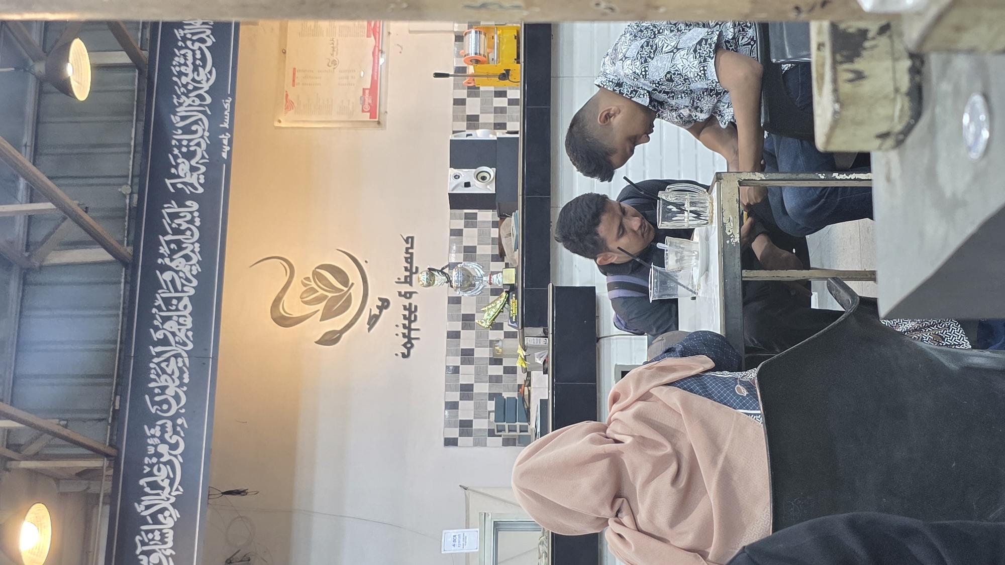 The scene appears to be inside a coffee shop named "Coffee & Beans", possibly located in Lhokseumawe, Indonesia, based on the Arabic script and the Indonesian phrase "ayat kunsi" visible in the image. The image is taken at an angle, looking down upon a group of people sitting at a table. Two men are leaning over the table and appear to be conversing. One man wears a blue-patterned shirt, and the other wears a striped shirt with a backpack. A woman in a peach-colored headscarf is also present at the table. Various glasses are visible on the table. The shop's interior features a tiled backsplash, an espresso machine, and decorative logos/lettering on the wall, along with lamps. The structural elements of the building's ceiling, including wooden beams, are visible.