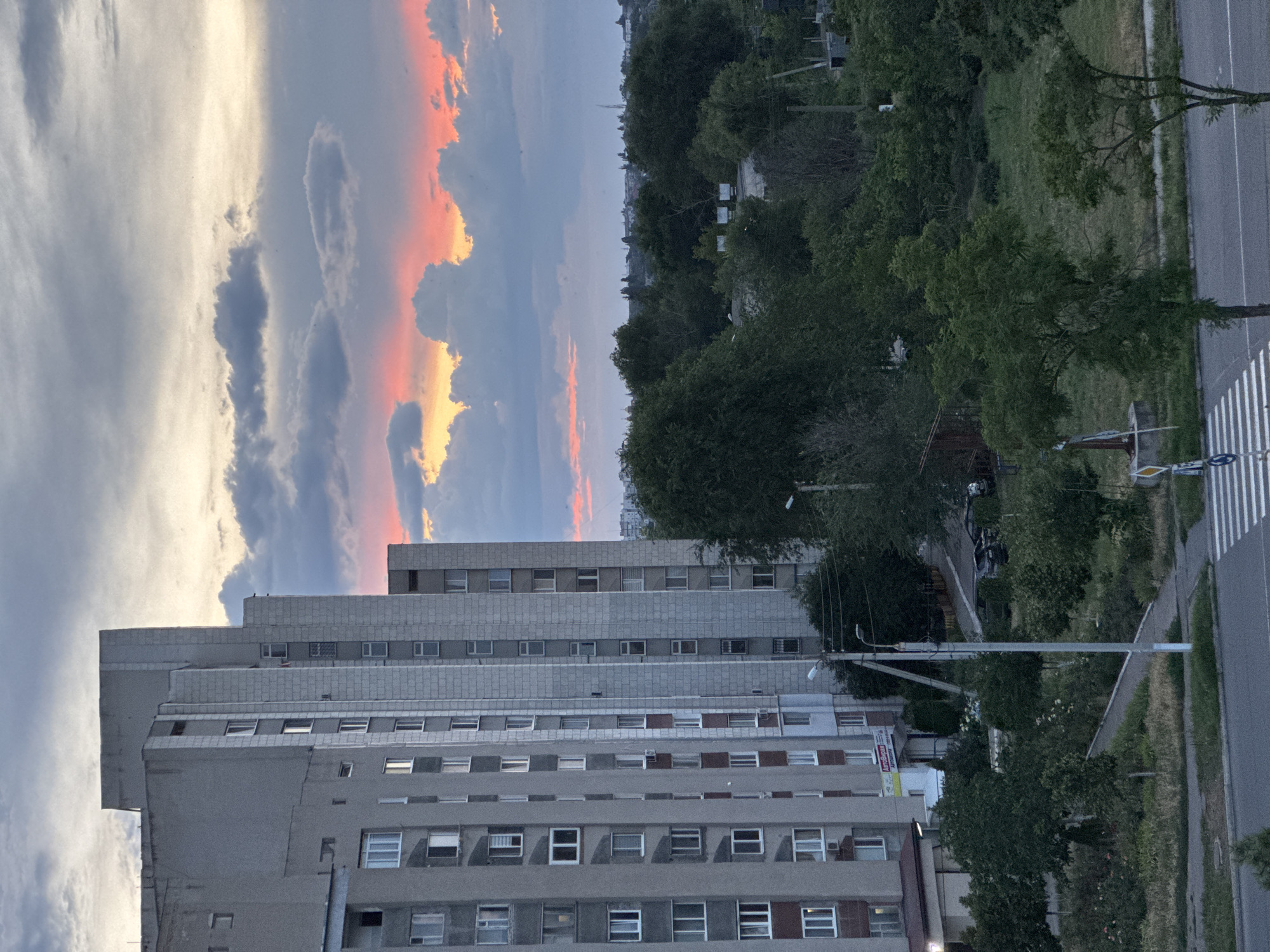 The image shows a wide-angle, high-angle view of a residential building and its surroundings in Tiraspol, Moldova, during sunset. The building, a tall, gray, multi-story structure with numerous windows, dominates the left side of the frame. Its architectural style appears to be Soviet-era. The sky above is filled with dramatic clouds, some tinged with the vibrant oranges and pinks of the setting sun, while others are darker and more ominous.
Below and to the right of the building, there is a dense area of green trees and vegetation, suggesting a park or natural area within the urban landscape. A road runs in the foreground, with a crosswalk clearly visible. A lamppost stands near the sidewalk in front of the building. In the background, more buildings and infrastructure are visible, hinting at the extent of the city.
There are no people or any discernable actions or interactions occurring in the scene. The focus is on the static elements of the urban environment and the atmospheric conditions of the sunset. The overall impression is one of a quiet evening in a residential part of Tiraspol.