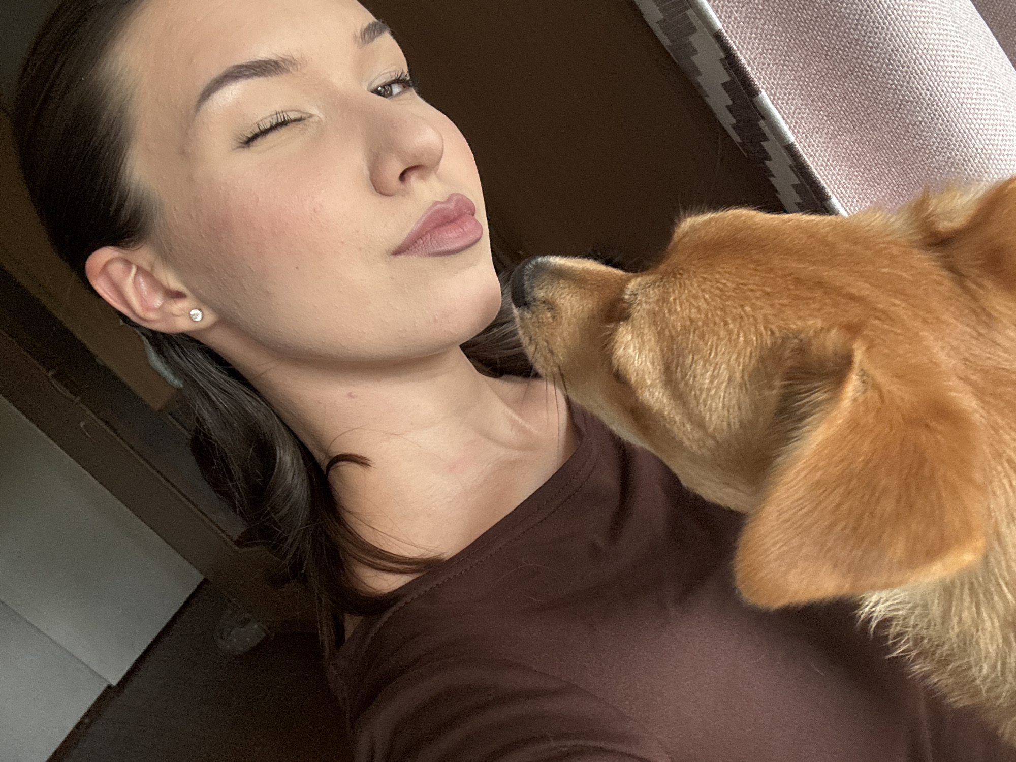 A young woman with dark hair, wearing a brown shirt and a small diamond earring, is captured in a close-up selfie. She is winking, and her lips are slightly parted. Her right cheek has a subtle pink blush. She is holding a small tan dog, which is looking up at her face. The background suggests an indoor setting, with hints of wood paneling or furniture, and a patterned fabric in the lower right corner. The lighting is soft, highlighting the woman's facial features and the dog's fur. There is no visible indication of the city of Inskoy, Russia.