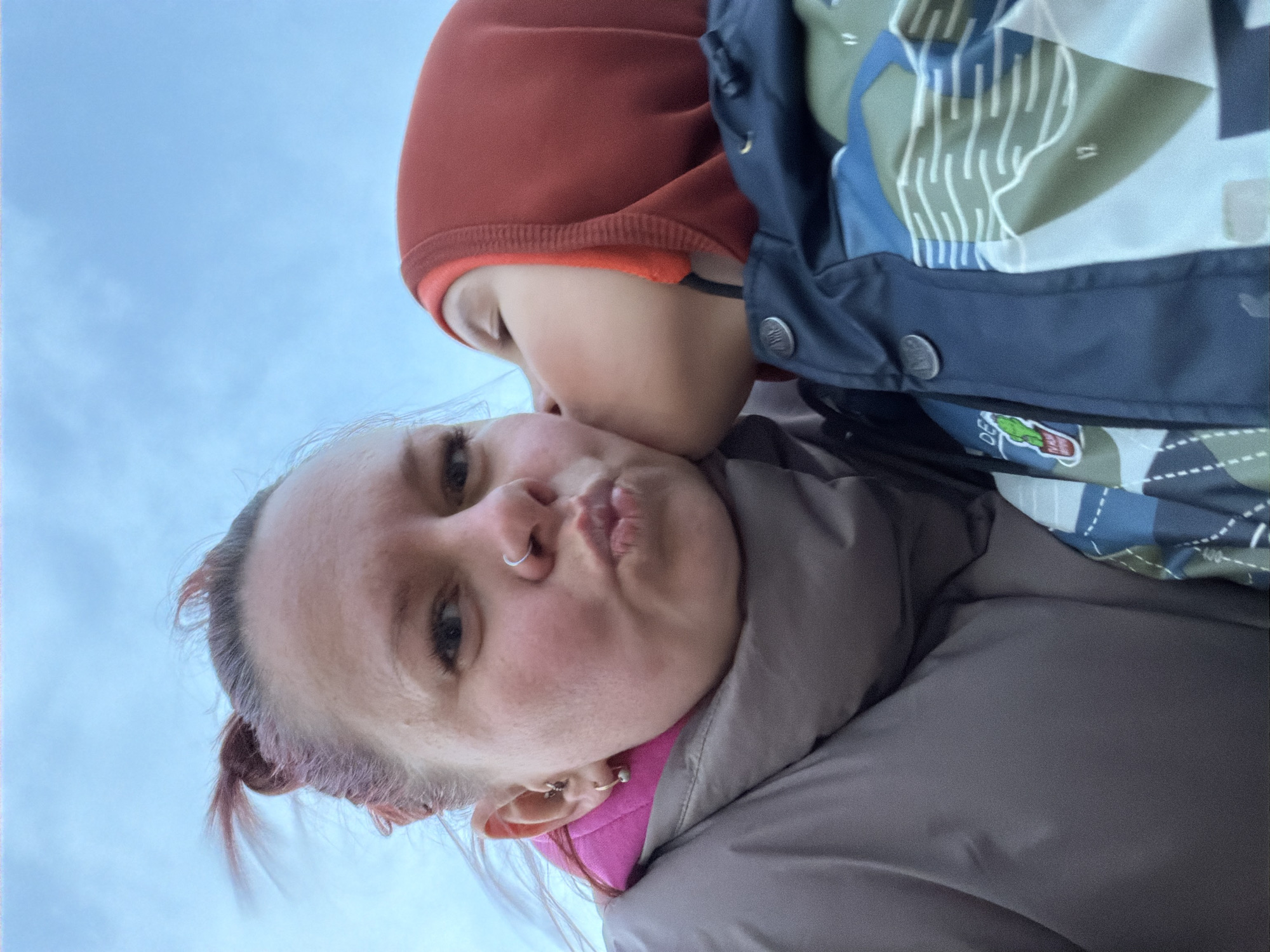 This upward-angled portrait, taken in Chaikovsky, Russia, features two individuals against a clear blue sky. In the foreground, an adult woman with light-colored hair, a nose ring, and multiple ear piercings looks directly at the camera with a playful pouting expression. She is dressed in a warm grey or brown puffer jacket layered over a bright pink garment. From behind and above, a child wearing a red hooded top and a patterned winter jacket with blue, green, and white abstract designs, gently kisses the woman's cheek. The child's jacket has a small green and white patch near the bottom, displaying the letters "DE" and what appears to be a small creature logo. The warm winter clothing of both subjects suggests a cool or cold outdoor temperature, characteristic of the location. The image conveys a tender and affectionate moment between the two, likely during the daytime under clear weather conditions.