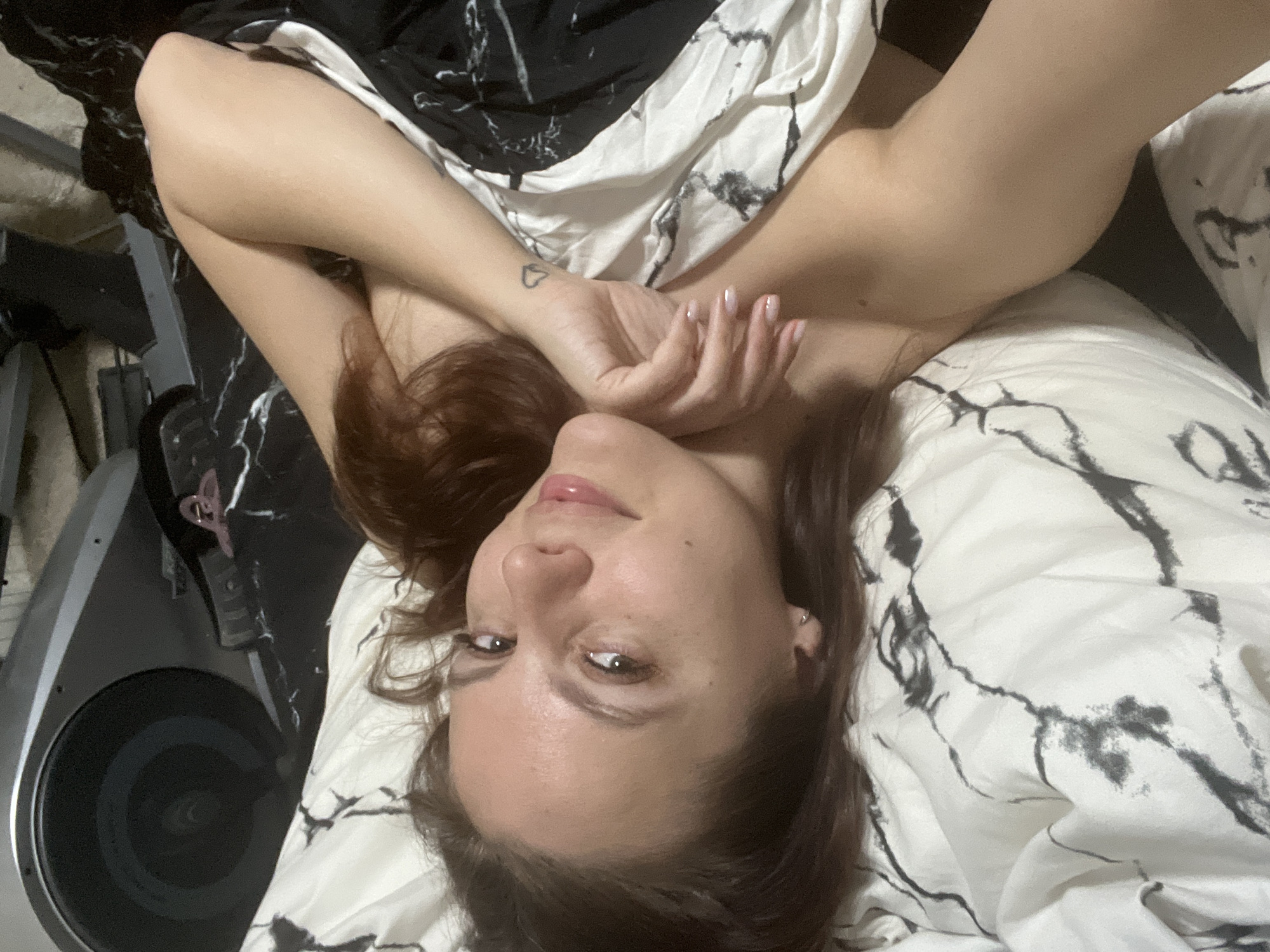 An image, rotated, depicts a woman with reddish-brown hair lying on a bed, looking directly upwards at the camera. Her head is positioned towards the bottom of the frame, and her body extends upwards. Her arms are crossed over her chest/stomach area, with her hands resting near her chin. A small, black outline of a heart is tattooed on her right wrist, and her fingernails are painted with a light, perhaps sheer pink, polish. She has an earring in her visible left ear. Her expression is neutral to slightly soft, with a hint of a gentle smile, and her lips appear glossy.
She is covered by bedding featuring a distinct black and white marble-like pattern, which also covers the surface she is lying on. In the background on the far left, partially visible, is a piece of exercise equipment, specifically the flywheel and housing of what appears to be an elliptical trainer or stationary bike. The setting is indoors, likely a bedroom, characterized by the relaxed posture and bedding. The lighting suggests daytime or bright artificial illumination, but no specific time or weather can be discerned. The overall scene conveys a personal and intimate moment, possibly in a home in Chaikovsky, Russia. There is no visible text in the image.