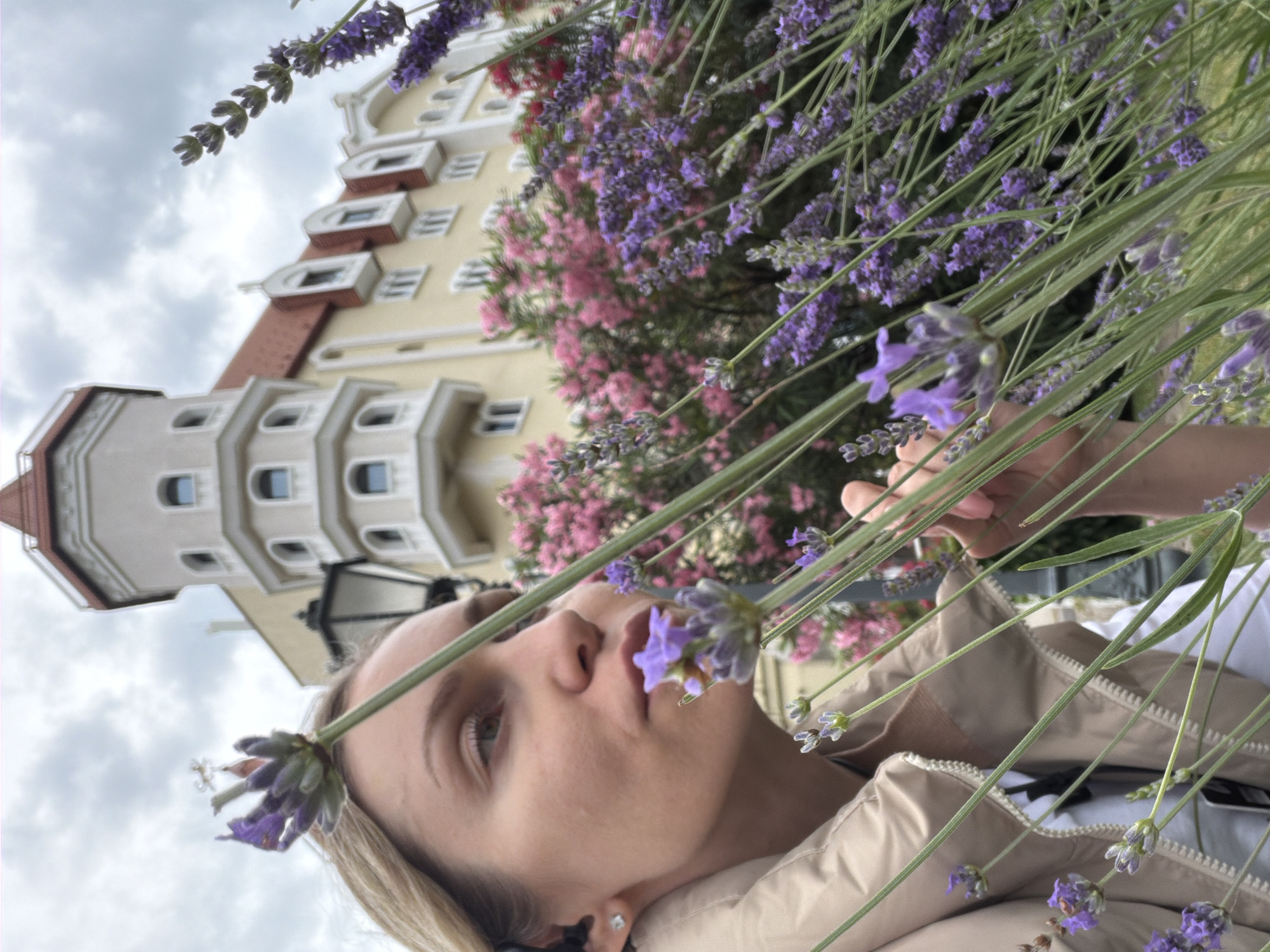 In the city of Sirius, Russia, a woman with blonde hair is captured in a medium shot, her face turned towards the left, her gaze directed slightly downward. She is wearing a beige jacket and a white top. Her right hand is partially visible, touching lavender flowers that are in the foreground, obscuring parts of her face and body. Behind her, a building with a light yellow facade and a red-tiled roof is visible, featuring arched windows and a prominent turret. Lush pink and red flowers are interspersed with the lavender, creating a vibrant floral backdrop. The sky above is overcast with clouds.