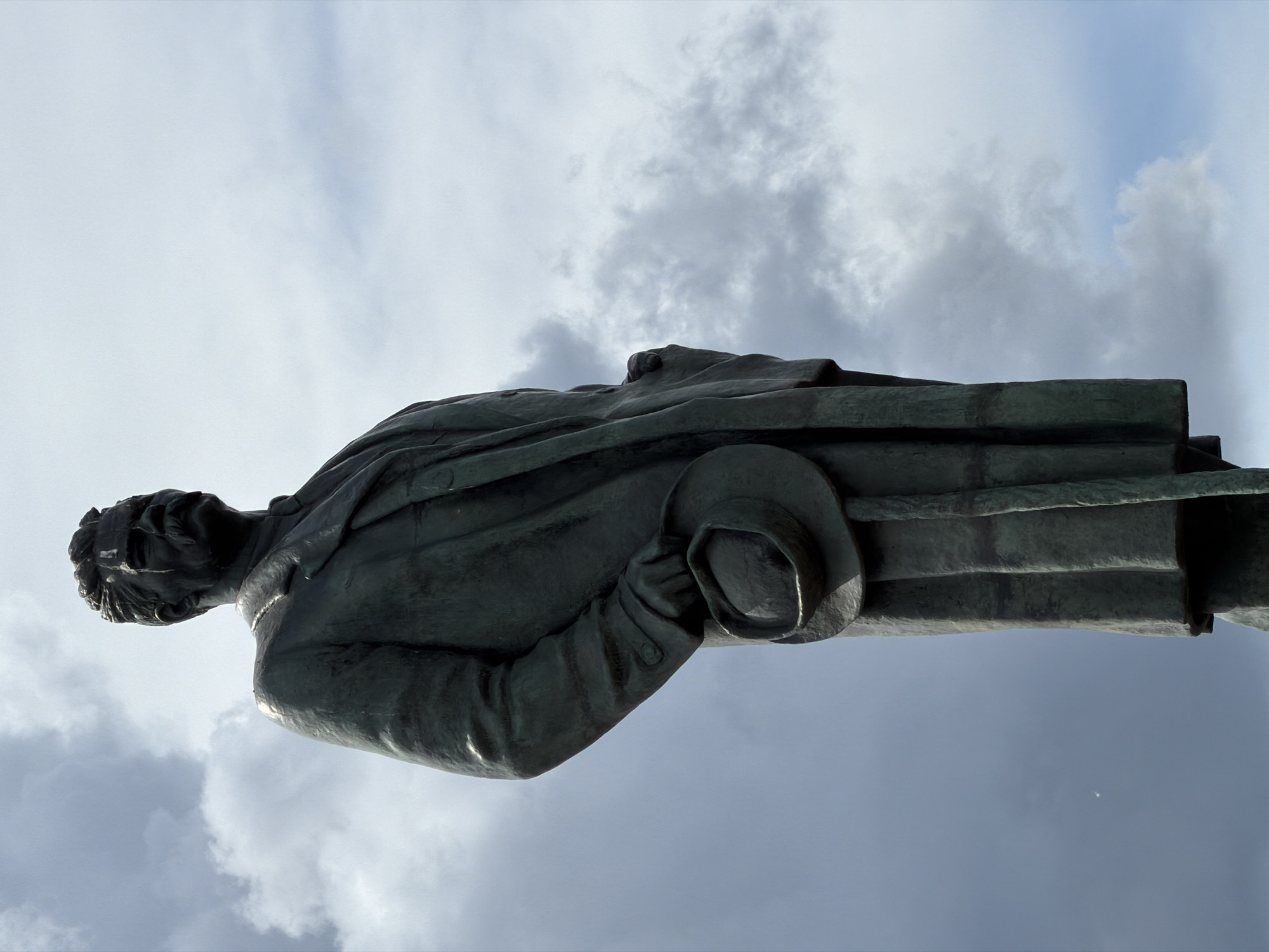 A low-angle shot shows a bronze statue of a man against a cloudy sky. The man is wearing a long coat and is holding a hat in his left hand. The statue is located in Moscow, Russia.