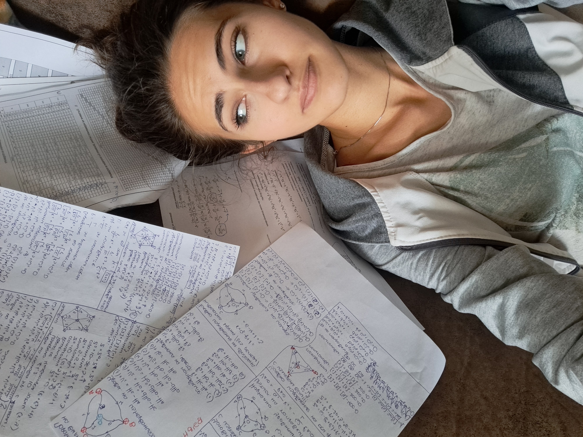 A young woman lies on a floor covered with papers containing mathematical equations and diagrams. She is wearing a gray and white hooded sweatshirt over a gray t-shirt, and a thin necklace. Her dark hair is pulled back, and she is looking upwards with a slightly quizzical expression. The location is not identifiable as Negovan, Bulgaria from the image alone. The scene depicts a state of being overwhelmed by academic work.