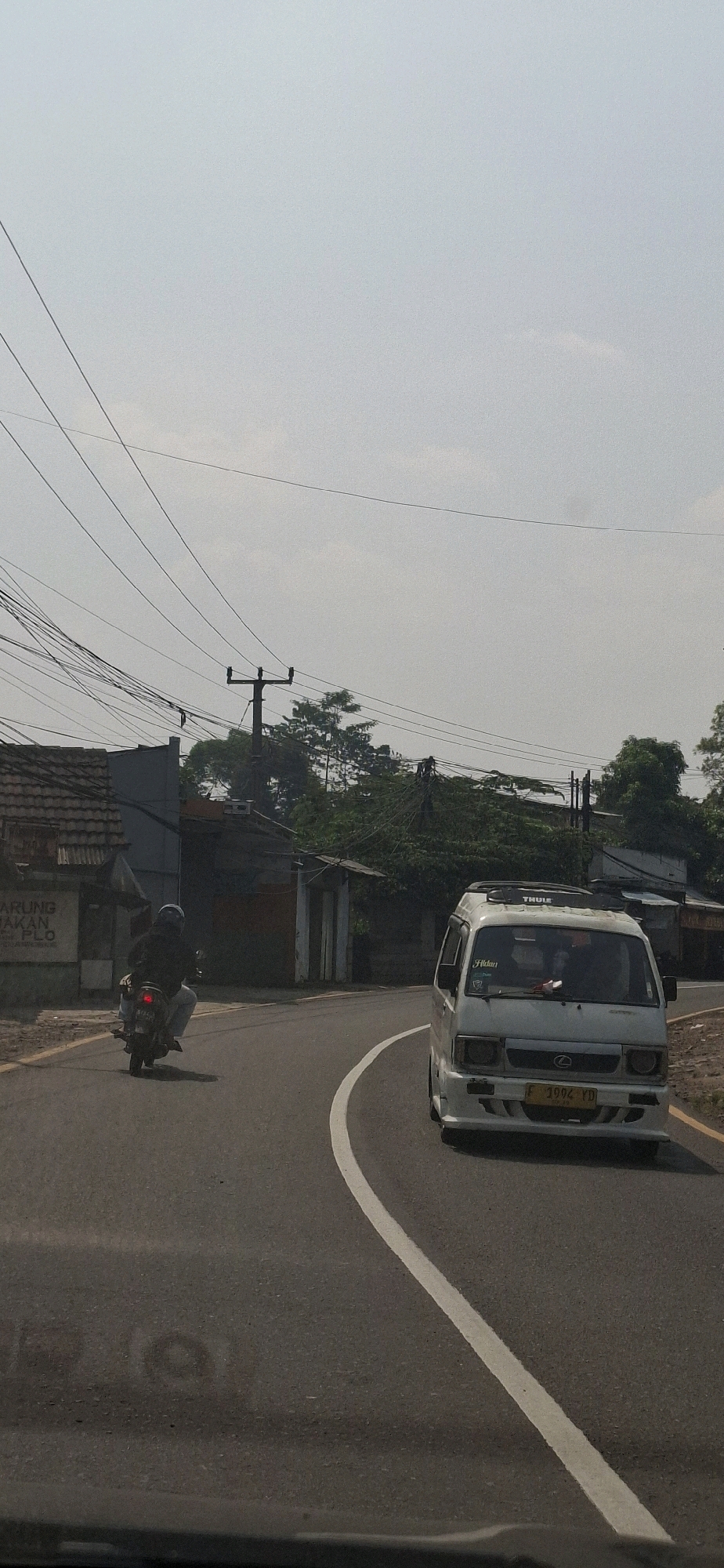 This is a vertical, eye-level, medium-wide shot taken on a road in Cilaku, Indonesia, on a hazy, bright day. The road curves to the right in the foreground, with a white line marking the edge. A white, boxy van with a black roof rack is traveling on the road, its front facing the viewer. The van has a yellow license plate that reads "F 1992 YD". To the left of the van, a person on a motorcycle is also traveling on the road, moving away from the viewer and towards the curve. The motorcyclist is wearing a dark helmet and dark clothing.
On the left side of the road, there is a small building with a tiled roof, and a sign that appears to have Indonesian text. Further back, there are more buildings and trees. Power lines are visible crisscrossing the sky. The environment appears to be a rural or semi-rural area with some signs of development. The hazy sky suggests it might be a warm, humid day.