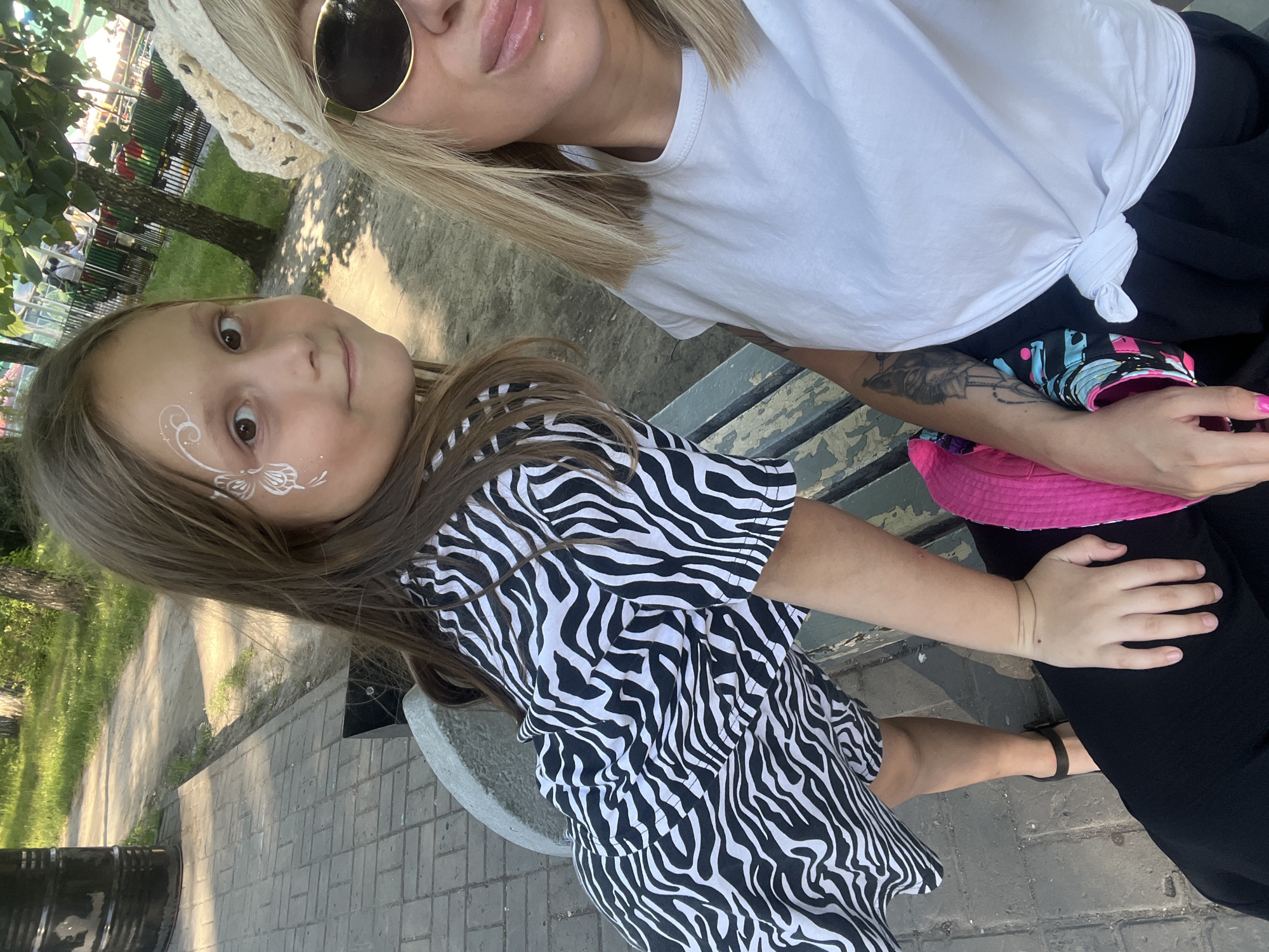 The media file shows two people, a woman and a young girl, sitting on a bench outdoors. The woman has blonde hair and is wearing sunglasses and a white t-shirt tied at the waist. She also has tattoos visible on her arm. The girl has brown hair with white face paint on her cheek that appears to be a butterfly. She is wearing a black and white zebra-print dress. A bright pink hat is visible on the bench. The scene appears to be a park or outdoor area with trees and a paved walkway. The lighting suggests it is daytime, likely sunny. The overall mood seems casual and relaxed.