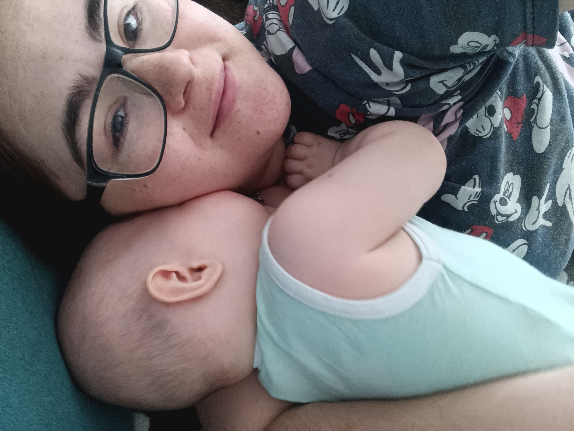 A woman wearing glasses and a blue Mickey Mouse patterned shirt is lying down with an infant. The infant is wearing a light blue undershirt with white trim. The woman's face is close to the infant's head. The background includes a teal-colored fabric. There is no indication of the city of Noviy, Russia in the image.