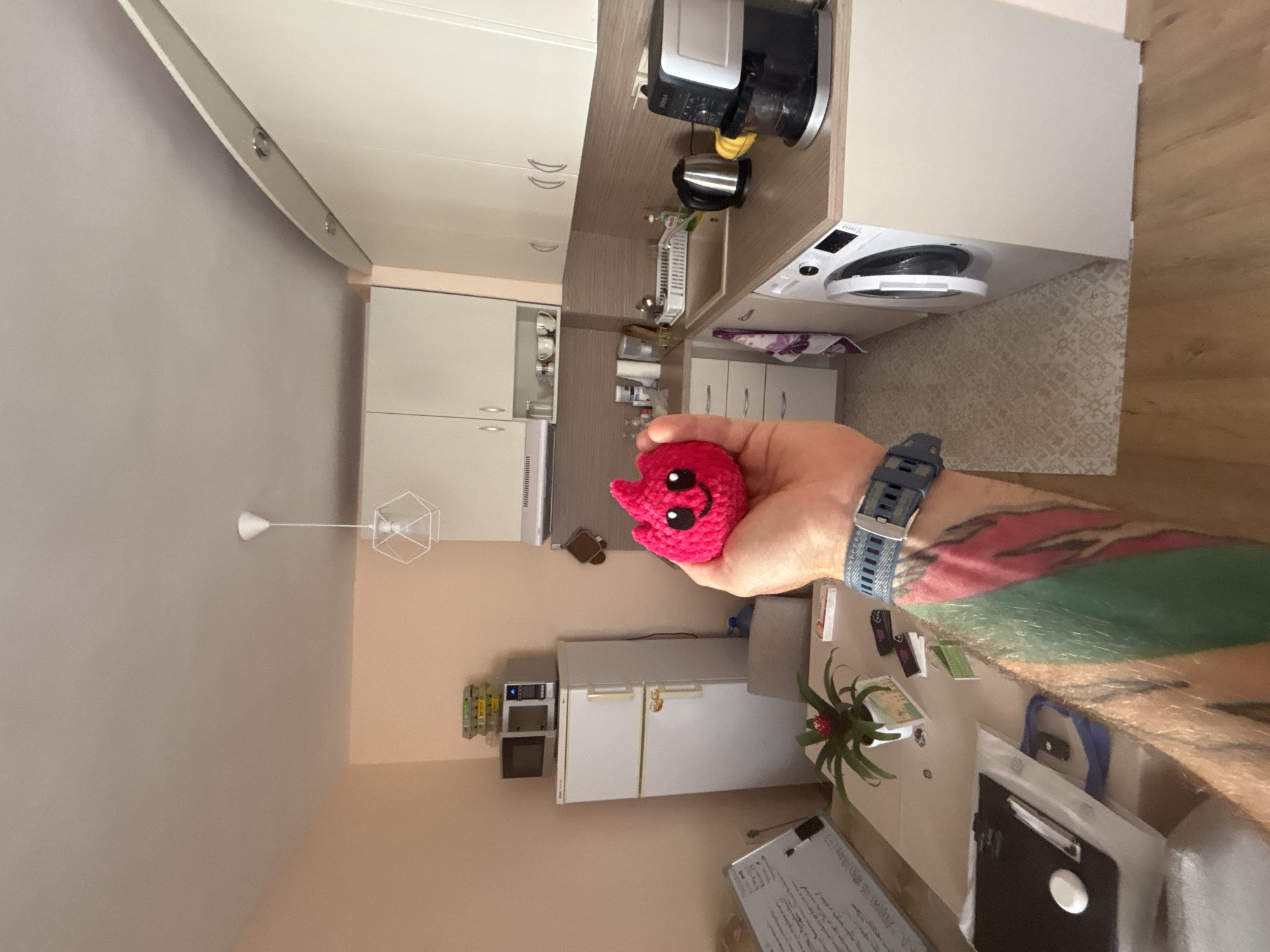 The image depicts a small kitchen area in an apartment. A person's hand, with colorful tattoos on their arm and wearing a watch, is holding a small, pink, crocheted stuffed toy that resembles a flame with a smiley face. The kitchen features white cabinets, a beige countertop, a sink, a washing machine, a coffee maker, a kettle, and a refrigerator with a microwave on top. A dining table is visible in the foreground with a plant and various items on it, including what appears to be a whiteboard or easel with writing. The flooring is a light-colored patterned tile. The apartment is likely located in the city of Varna, Bulgaria.