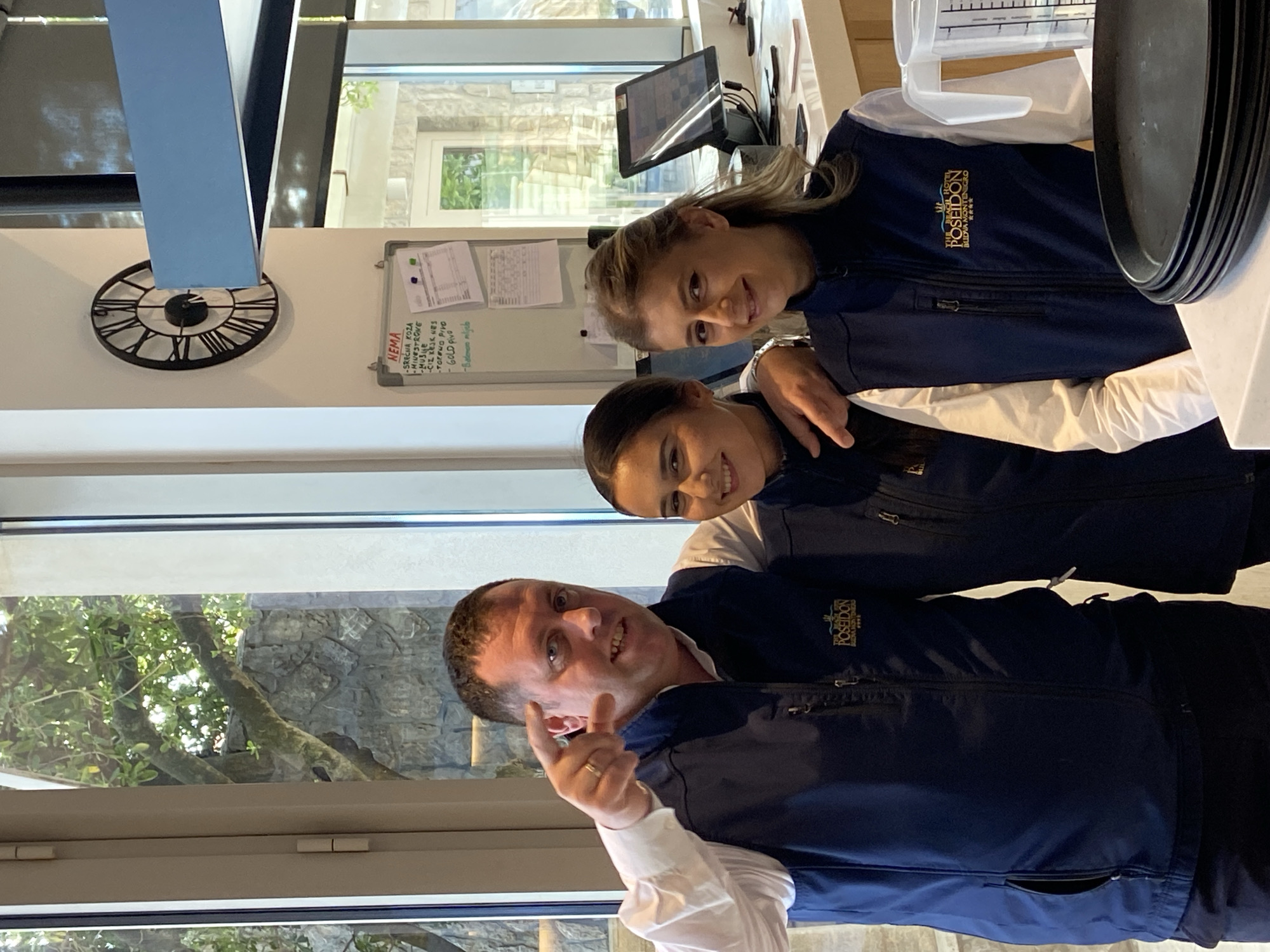 The image shows three people smiling and posing for a photo indoors. They are all wearing navy blue vests with the word "POSEIDON" and "BUDVA MONTENEGRO" embroidered on them. The man in the front is pointing his finger, and the women are positioned behind him. In the background, there's a window showing trees and a stone wall. To the left, there's a decorative clock on the wall. A whiteboard with a list of items, including "SRECHA KOZA," "MINESTRONE," "CIZ KEJK HES," and "GOLD pivo," is visible. A computer screen and some kitchen equipment are also partially visible. The location seems to be an indoor setting possibly connected to the Poseidon brand, with visible outdoor scenery through the window. The precise location within Prijevor cannot be determined from the image alone.