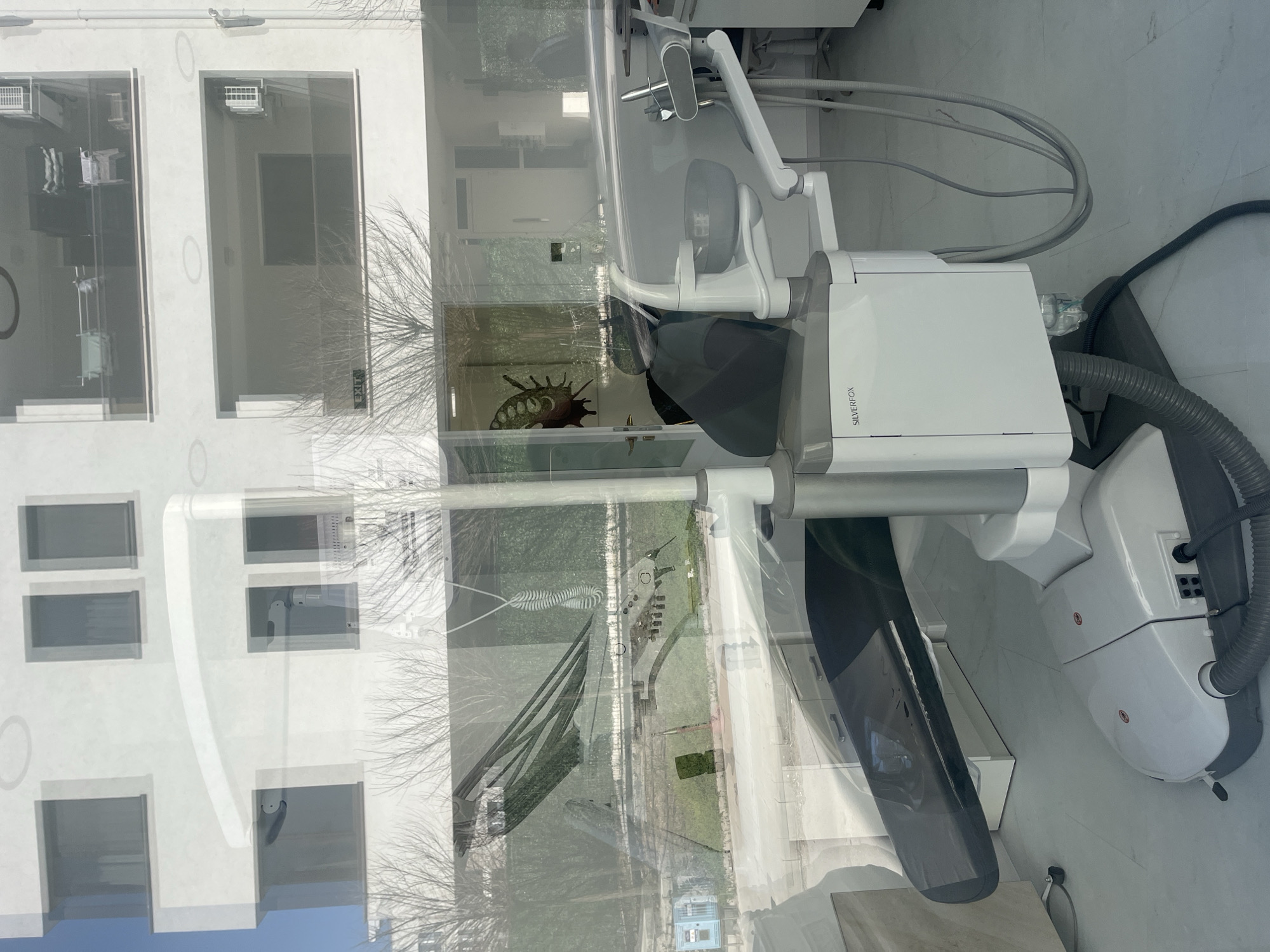 The image shows a dental office interior as seen through a window, with reflections of a building exterior. The dental equipment is modern, with a white chair, overhead light, and various hoses and attachments. The name "SILVERFOX" is visible on the equipment. A decorative shell is on display in the background of the office. The exterior reflection shows a white building with several rectangular windows. The reflections are blurred by the window glass. I have no information about the specific city in Bulgaria shown in the picture.