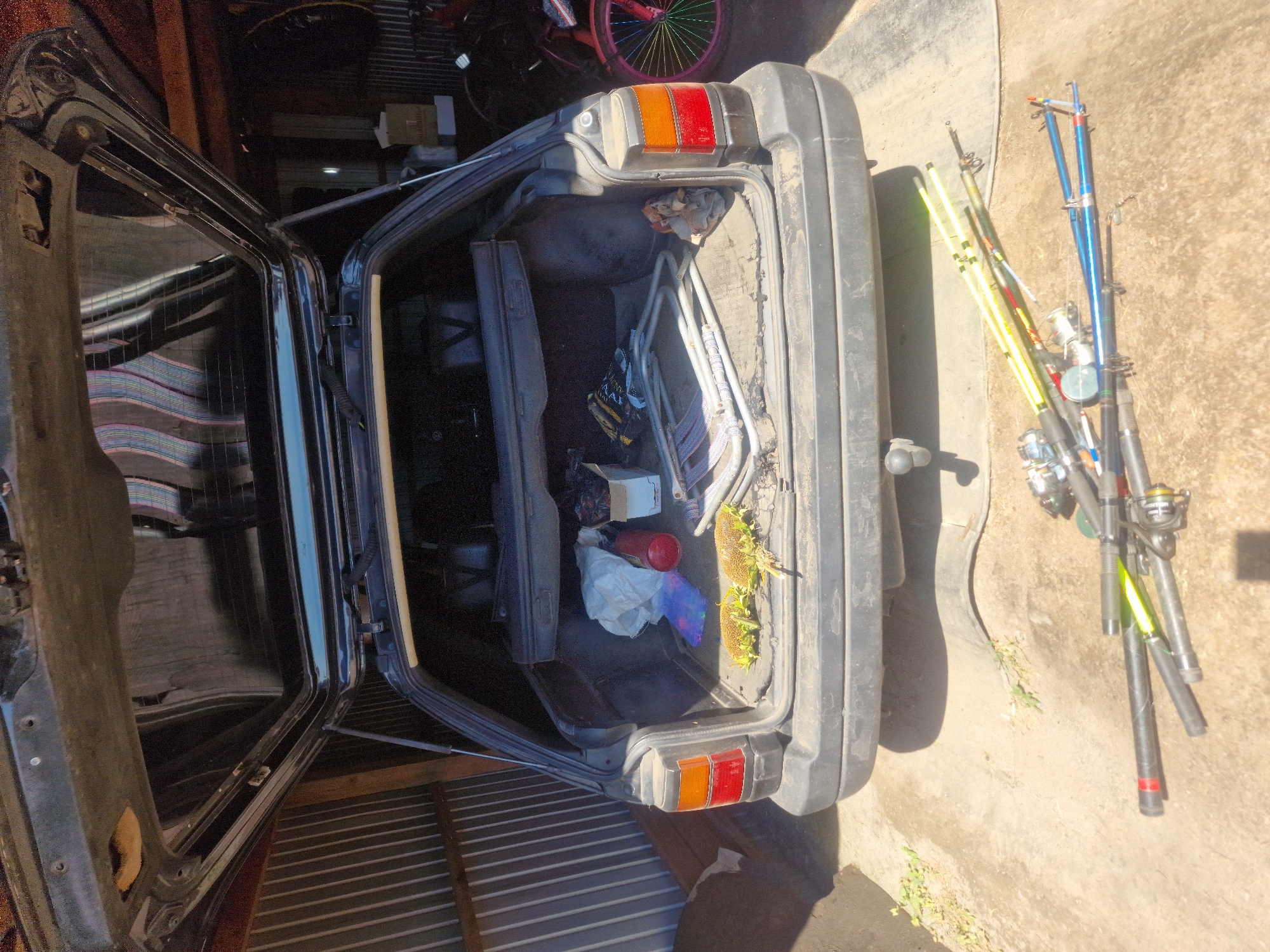 The image shows the open trunk of a black station wagon parked outdoors on a dirt and concrete surface, likely in Makinsk, Kazakhstan. The trunk contains several items, including two dried sunflowers, a red fire extinguisher, a white plastic bag, a box, and some metal folding chairs. Lying on the ground in front of the car are several fishing rods and reels. In the background, a corrugated metal wall is visible, as is a bicycle with brightly colored wheels. The overall impression is one of domestic storage and outdoor recreational equipment.
