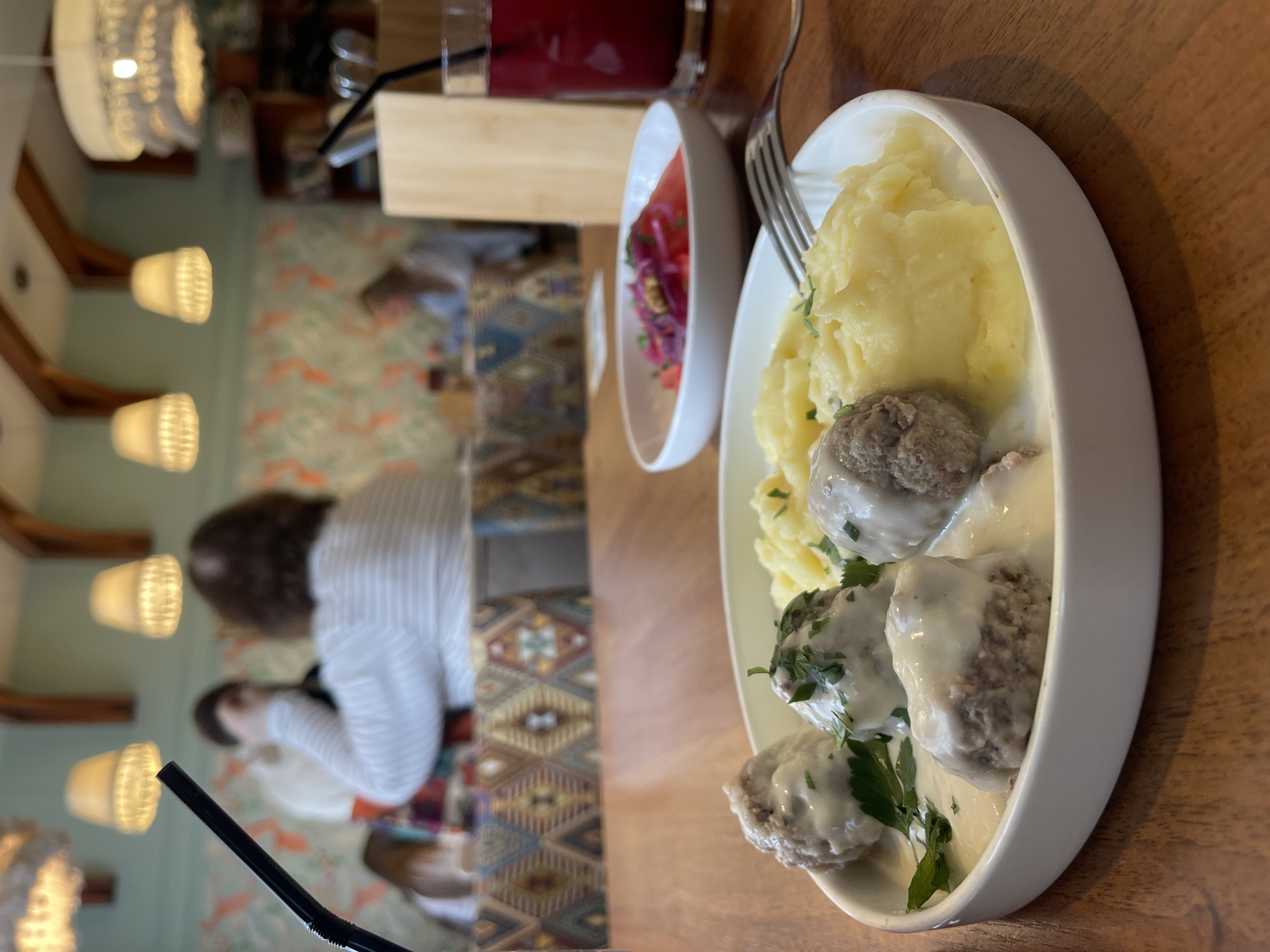 A close-up, top-down shot shows a meal of meatballs in cream sauce with mashed potatoes on a wooden table. The plate is white and oval-shaped. There are at least four meatballs visible, generously covered in a creamy white sauce and sprinkled with fresh parsley. The mashed potatoes are a vibrant yellow and appear smooth and fluffy. A fork rests on the edge of the plate. To the left of the plate, a small white bowl contains a salad with sliced tomatoes and red onions. In the background, a glass of dark red liquid, possibly juice or a beverage, is visible. Further to the left, several decorative lamps with a woven texture hang from the ceiling. The room's wallpaper has a repeating pattern of orange and white flora. Two individuals are partially visible in the background, blurred due to the shallow depth of field. One person, wearing a striped shirt, has curly brown hair. Another person is wearing a white shirt. The scene appears to be set indoors, possibly in a restaurant in Izhevsk, Russia, given the context. The lighting suggests it could be daytime, but the indoor setting makes it difficult to determine the exact time of day or weather conditions.