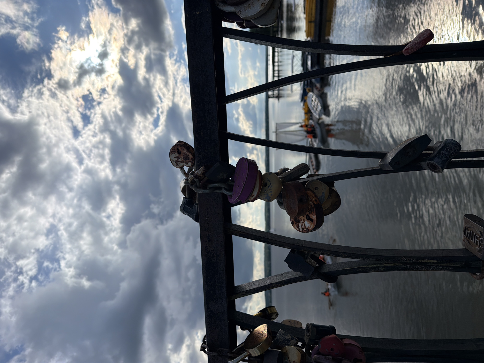 A bridge in Votkinsk, Russia, is adorned with "love locks." These padlocks, attached to the bridge's metal railing, symbolize enduring love. The locks are of various sizes, colors, and materials. The background features a body of water, likely a lake or river, under a cloudy sky. A boat can be seen in the distance.
