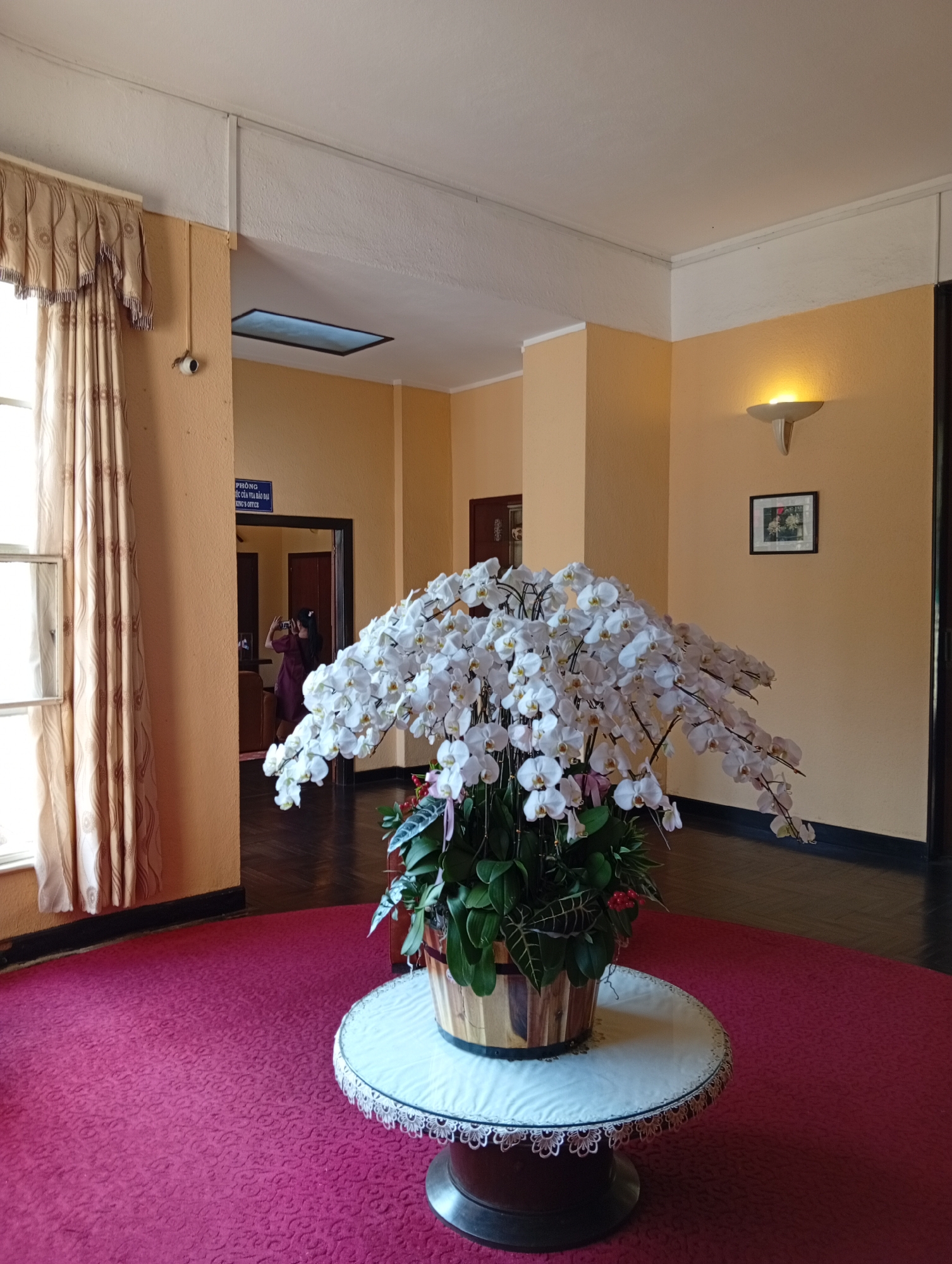 The photograph shows a large arrangement of white orchids in a woven pot, situated on a round table covered with a white cloth and lace trim. The table sits on a red patterned carpet that covers a significant portion of the floor. The background reveals a room with walls painted in a warm, light-orange hue. A doorway leads to another room, where a person is seen holding a phone, seemingly taking a picture. A sign above the doorway contains Vietnamese script, along with the English words "KING'S OFFICE." A window is to the left of the doorway. The architectural details, wall color, artwork, and specific signage suggest the location is likely within a historical building or landmark in the city of Da Lat, Vietnam. A wall sconce provides illumination on the right.