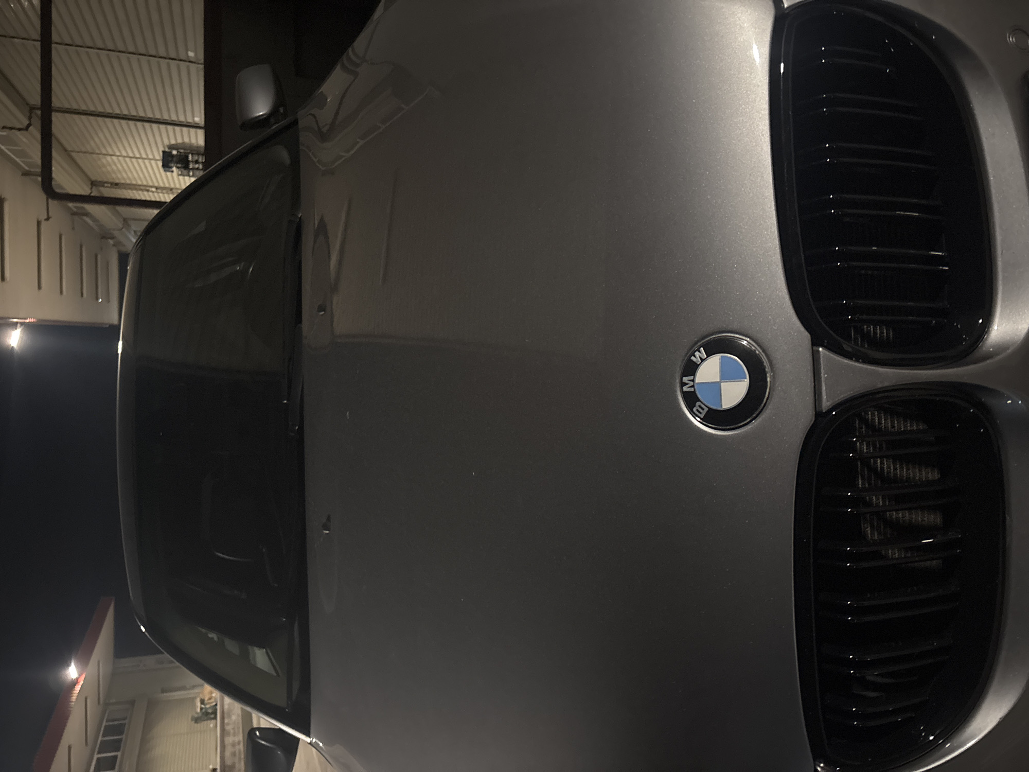 The image displays a close-up, frontal view of a metallic gray BMW sedan under artificial lighting, likely at night or in an enclosed facility. The vehicle's hood, dark windshield, and prominent dual kidney grilles with black vertical slats are central. The BMW emblem, featuring its blue, white, and black circular design with "BMW" text, is clearly visible on the hood. Overhead light sources are reflected on the car's hood and windshield.
The background on the left reveals a building structure with a red-trimmed awning or roofline and horizontal window or blind features below it, illuminated by a bright light. On the right, another section of a building or an adjacent structure is visible, characterized by multiple horizontal ceiling lights and walls with a corrugated or roll-up door appearance. A dark, industrial-style metal frame and a piece of equipment are present in the right background. No individuals are depicted in the scene. This scene is situated within the city of Plovdiv, Bulgaria.