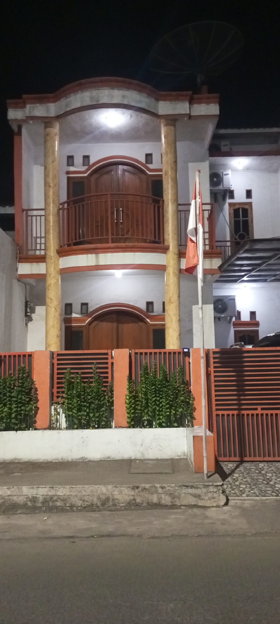 The image depicts the front of a two-story house at night. The house has a white facade with orange accents and features a prominent balcony with wooden railings on the second floor. Two large, arched wooden doors are visible on both the ground and second floors, with smaller square windows flanking them. The house is supported by two large, cream-colored columns with marble-like veining. A satellite dish is mounted on the roof.
In front of the house, an orange metal fence runs horizontally, with sections of vertical slats and solid panels. Lush green plants are planted in front of the fence. A flagpole with the Indonesian flag (red and white) is positioned to the right of the entrance. Two outdoor air conditioning units are mounted on the exterior walls of the house. The ground in front of the fence is paved with cobblestones, and a sidewalk separates it from the asphalt road. The scene appears to be located in the city of Setu, Indonesia.