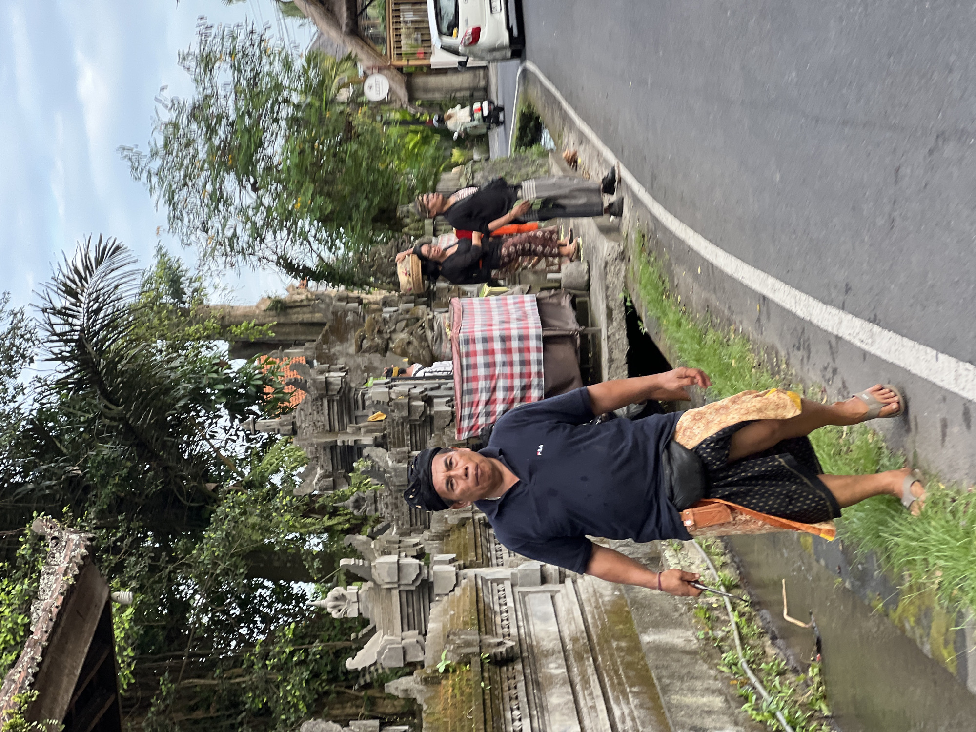 The image shows a man standing on a sidewalk in Ubud District, Indonesia. He is wearing a dark polo shirt, a black headdress, and a patterned sarong, and is holding a metal tool. Behind him is a Balinese temple structure with detailed stone carvings and greenery. Two other individuals are on the sidewalk further back, one carrying a basket on their head. A road runs alongside the sidewalk, with a white car and scooter visible in the background. The sky is overcast, and the overall scene suggests a cultural or ceremonial setting.