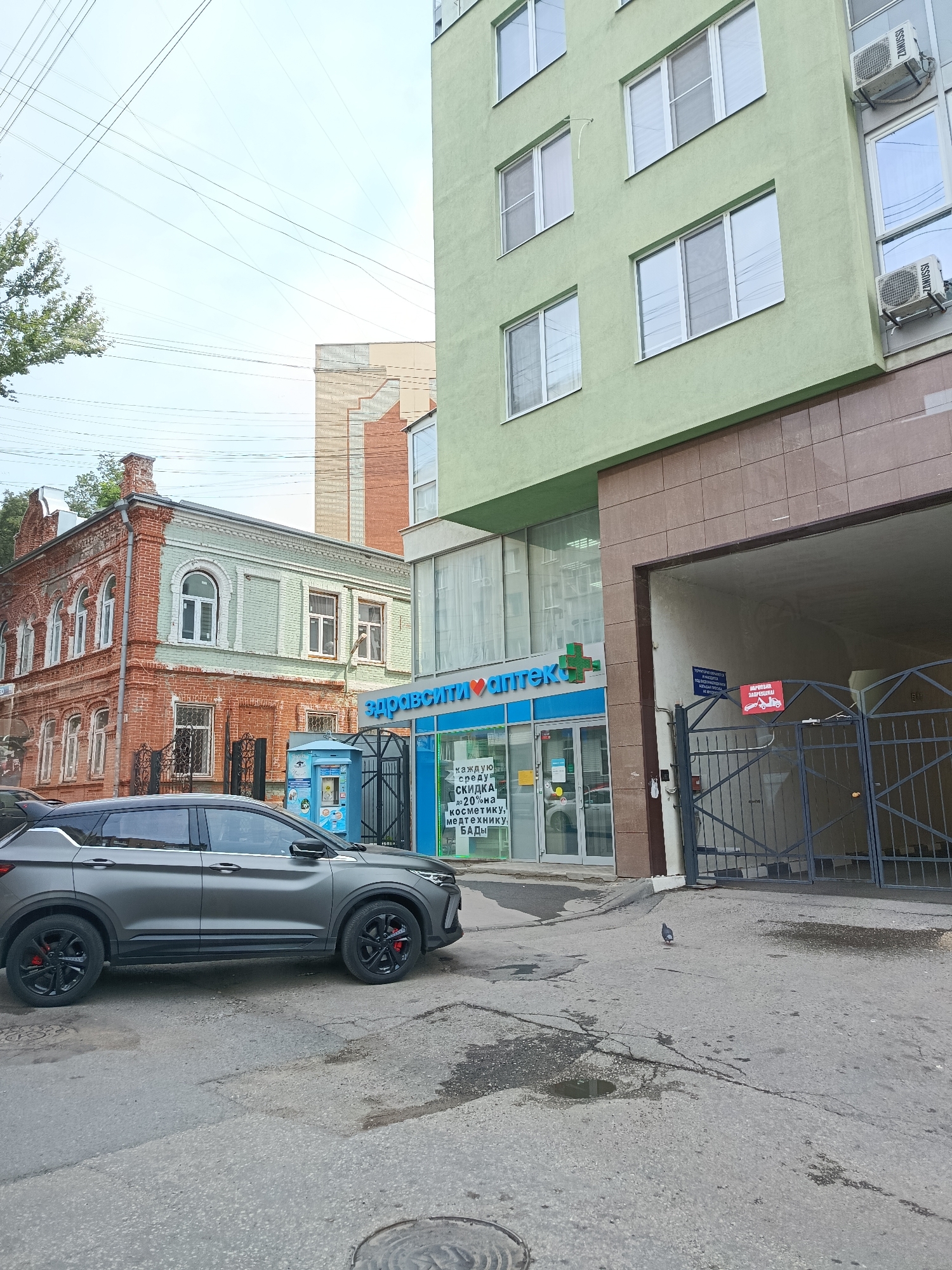 A gray SUV is parked on a paved street in front of a pharmacy with a green and blue sign that reads "Здравсити АПТЕК". The pharmacy is located in a modern building with light green walls. To the left of the modern building, there is an older red brick building with arched windows. The sky is overcast. A pigeon is walking on the street in front of the pharmacy. There is a metal gate to the right of the pharmacy, leading into a covered area. A small red sign with a tow truck symbol is visible on the gate. There are several power lines visible in the sky.