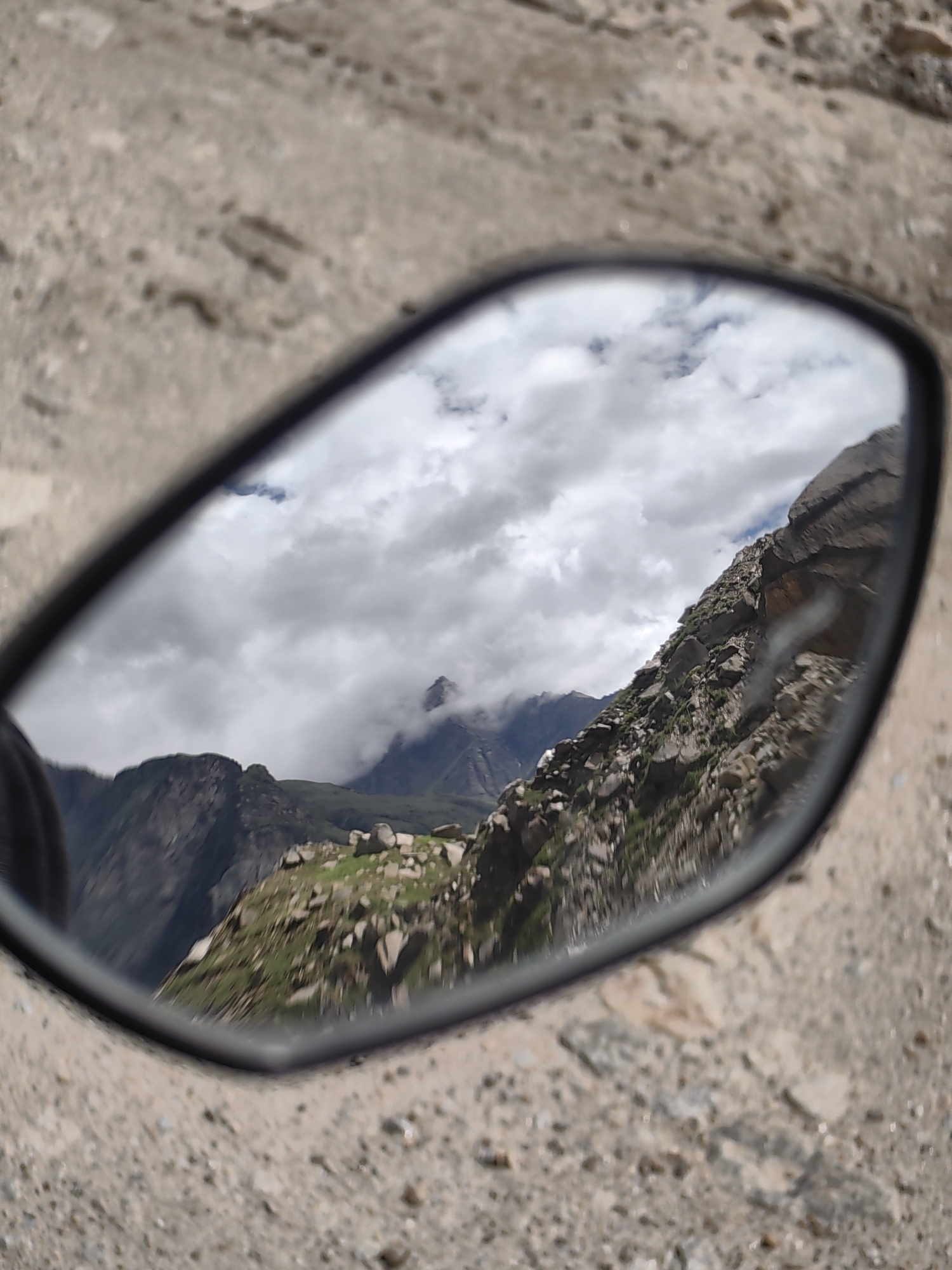 The image shows a motorcycle side-view mirror reflecting a mountainous landscape. The reflection shows rocky, barren slopes with patches of green vegetation. The sky is filled with dramatic, cloudy formations, with some mountains peeking through the clouds. The ground outside the mirror is a dusty, rocky terrain. The location is identified as Burwa, India. There are no people or animals visible in the image.