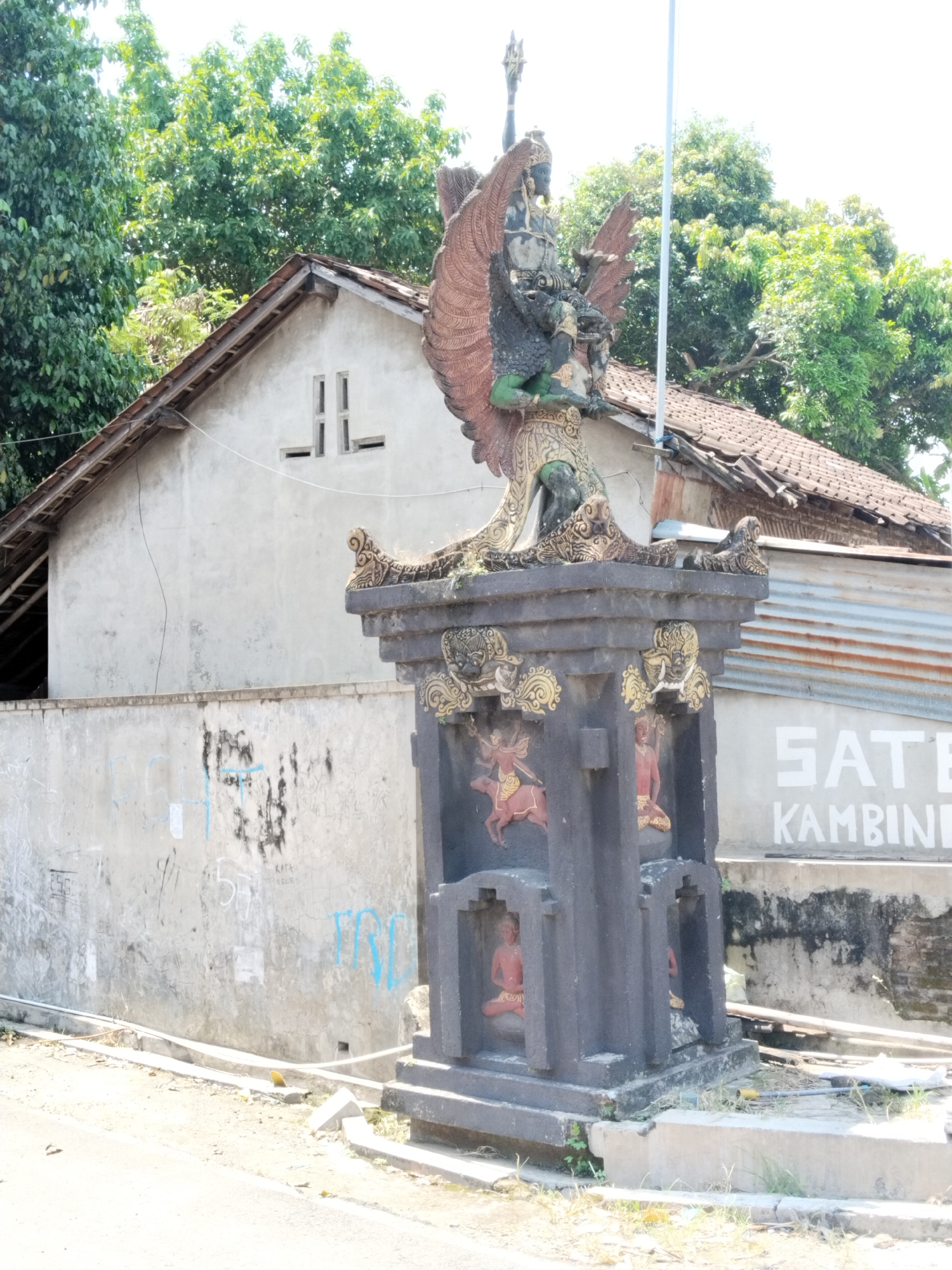 A color, eye-level, medium shot depicts an outdoor scene in Masaran, Indonesia. The statue of Garuda sits atop a square pillar, the pillar adorned with figures on each side. Behind the statue, a gray, single-story building with a tiled roof is visible. To the left, a gray wall is marked with faded blue graffiti. To the right, another building with a metal corrugated wall is partially visible, with the word "SATE" and the beginning of "KAMBING" painted on it. The foreground is a mix of dirt and concrete, suggesting a roadside or street view. Lush green trees fill the upper part of the image, suggesting a tropical or subtropical environment. The scene is brightly lit, indicating daylight.
