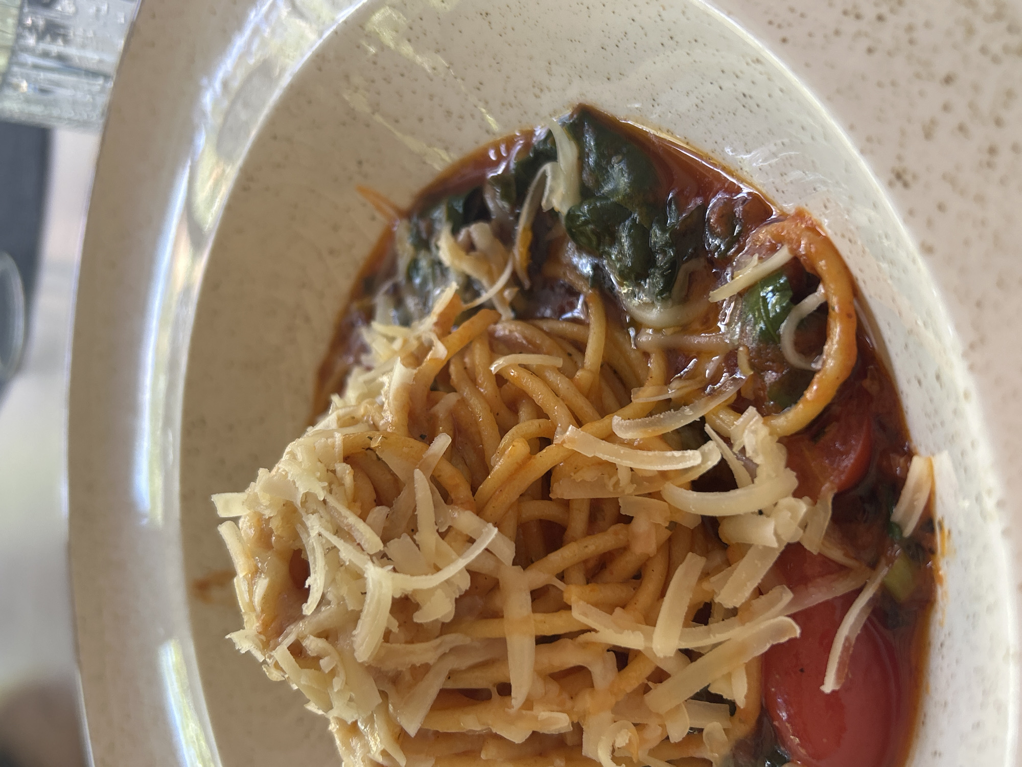 A close-up, overhead shot captures a shallow bowl of spaghetti topped with shredded cheese, tomato sauce, and visible pieces of spinach and tomato. The bowl itself is speckled with a light beige color and rests on a white surface. The lighting suggests an indoor setting, possibly a restaurant, with hints of natural light coming from the top left. No people or specific landmarks are visible in the image. The image is taken from an upward angle, focusing on the food. No information about the city of Sofia, Bulgaria, can be inferred from this image.