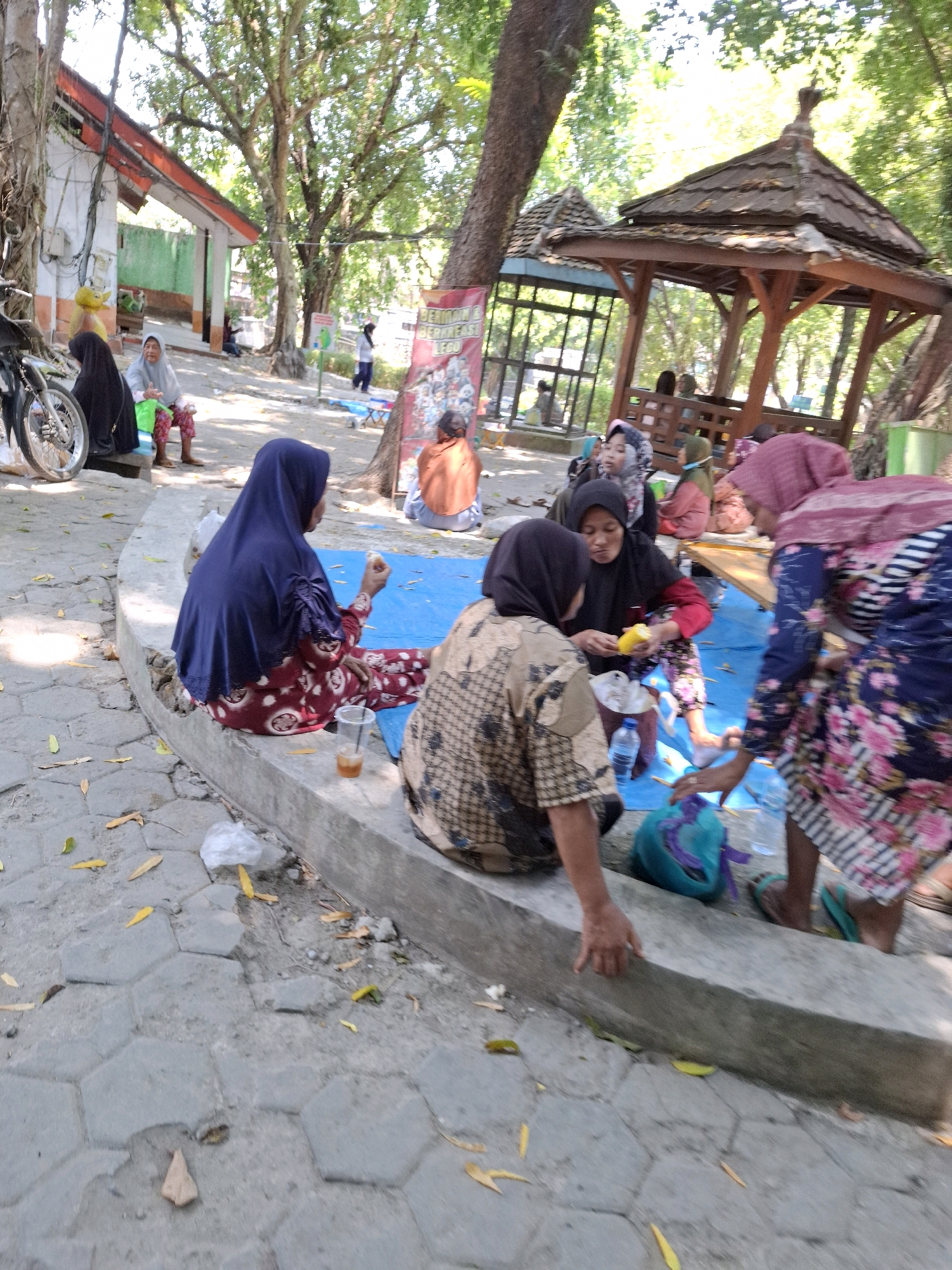 A group of people are gathered on a blue tarp on the ground, some sitting on a concrete ledge. They are outdoors in a shaded area with trees and a wooden gazebo visible in the background. A motorcycle is parked to the left, and a building with a red roof is also in the scene. The people are dressed in casual clothing, many wearing headscarves. Some individuals appear to be eating or sharing food. The ground is paved with hexagonal stones, and there are fallen leaves scattered around. A banner with text is visible in the background. The scene appears to be a casual gathering in a public park or recreational area in Ngawi, Indonesia.