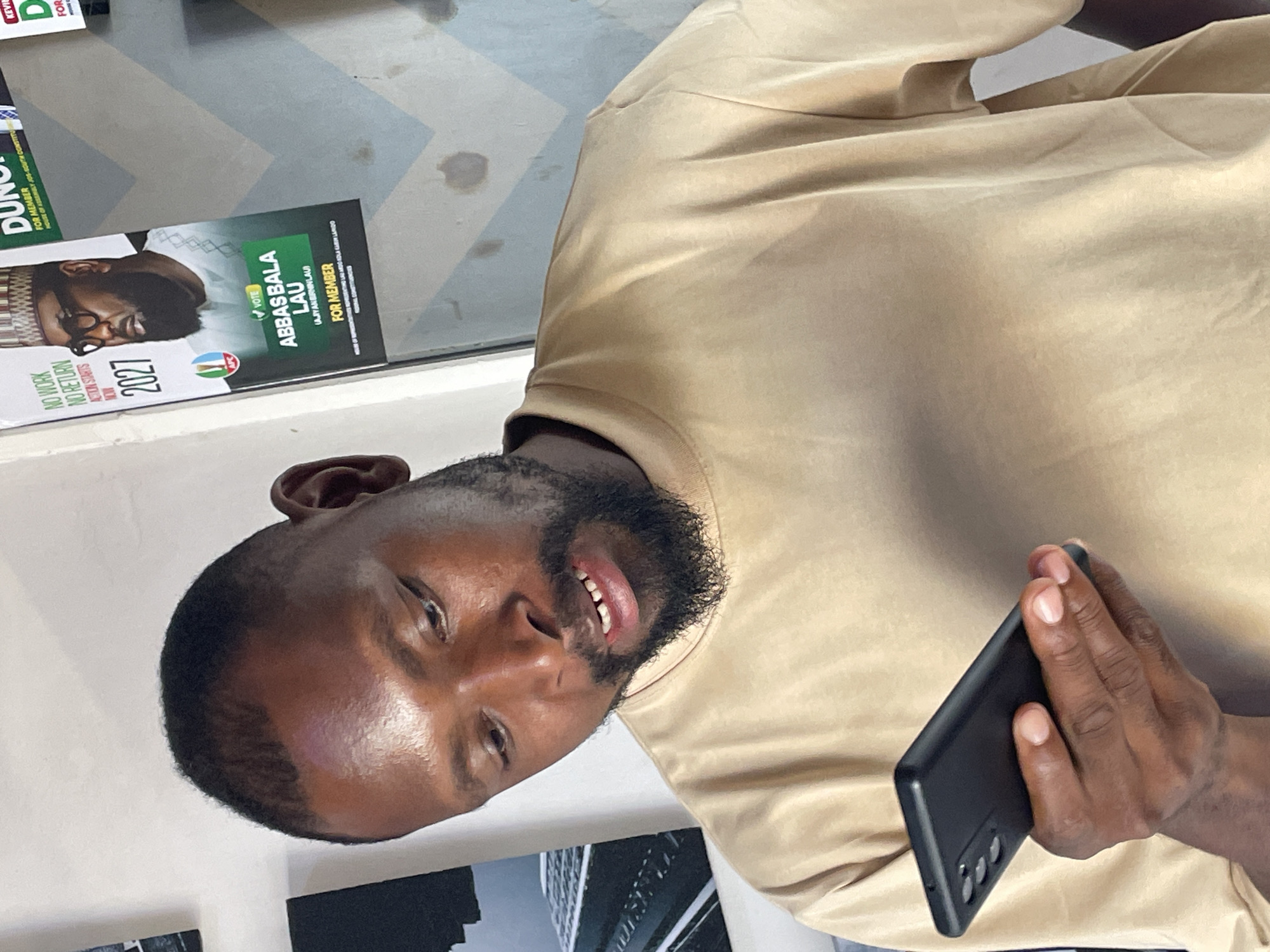 The image shows a man holding a black smartphone and looking down at it. He has dark skin, short hair, and a beard. He is wearing a tan t-shirt. In the background, there is a poster for "ABBAS BALA LAU FOR MEMBER". The poster features a man wearing glasses and a cap, with text that includes "NO WORK NO RETURN" and "ACTION STARTS NOW 2027". The setting appears to be indoors, with a white wall and a patterned section of the wall visible. Based on the presence of the political poster, the image was likely taken in Jos, Nigeria, during an election campaign period.