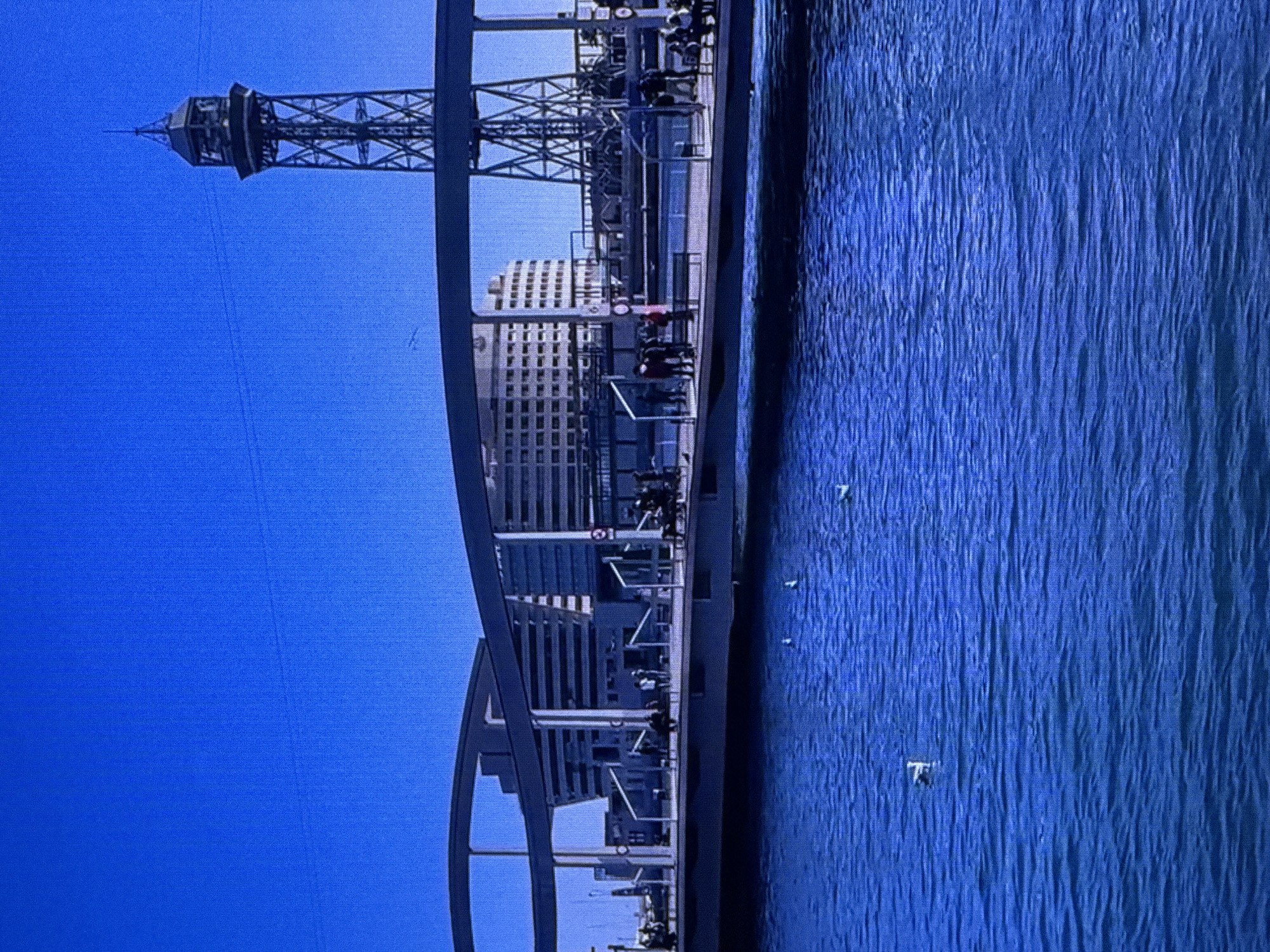 This is a vertical, cropped image of a modern cityscape with a prominent tower and water. The scene is set against a bright, clear blue sky, suggesting a sunny day. The water is a vibrant blue and appears calm with gentle ripples.
In the foreground, a portion of a modern architectural structure, possibly a bridge or walkway, is visible. It has a sleek, metallic design with curved supports. Along this structure, several people are walking and standing, appearing as small silhouettes against the bright background. They seem to be enjoying a leisurely stroll or observing the surroundings.
Dominating the middle ground is a tall, lattice-like tower with a distinctive observation deck or structure at its peak. Behind the tower, a modern, multi-story building with numerous windows can be seen, indicating an urban environment. The building's architecture is contemporary and geometric.
The image captures a vibrant and active urban scene. The bright sky and clear water suggest daytime. While specific emotions are hard to discern from the silhouettes, the overall impression is one of a pleasant, perhaps tourist-oriented location. There is no visible text within the image. The scene appears to be a waterfront area in a modern city.