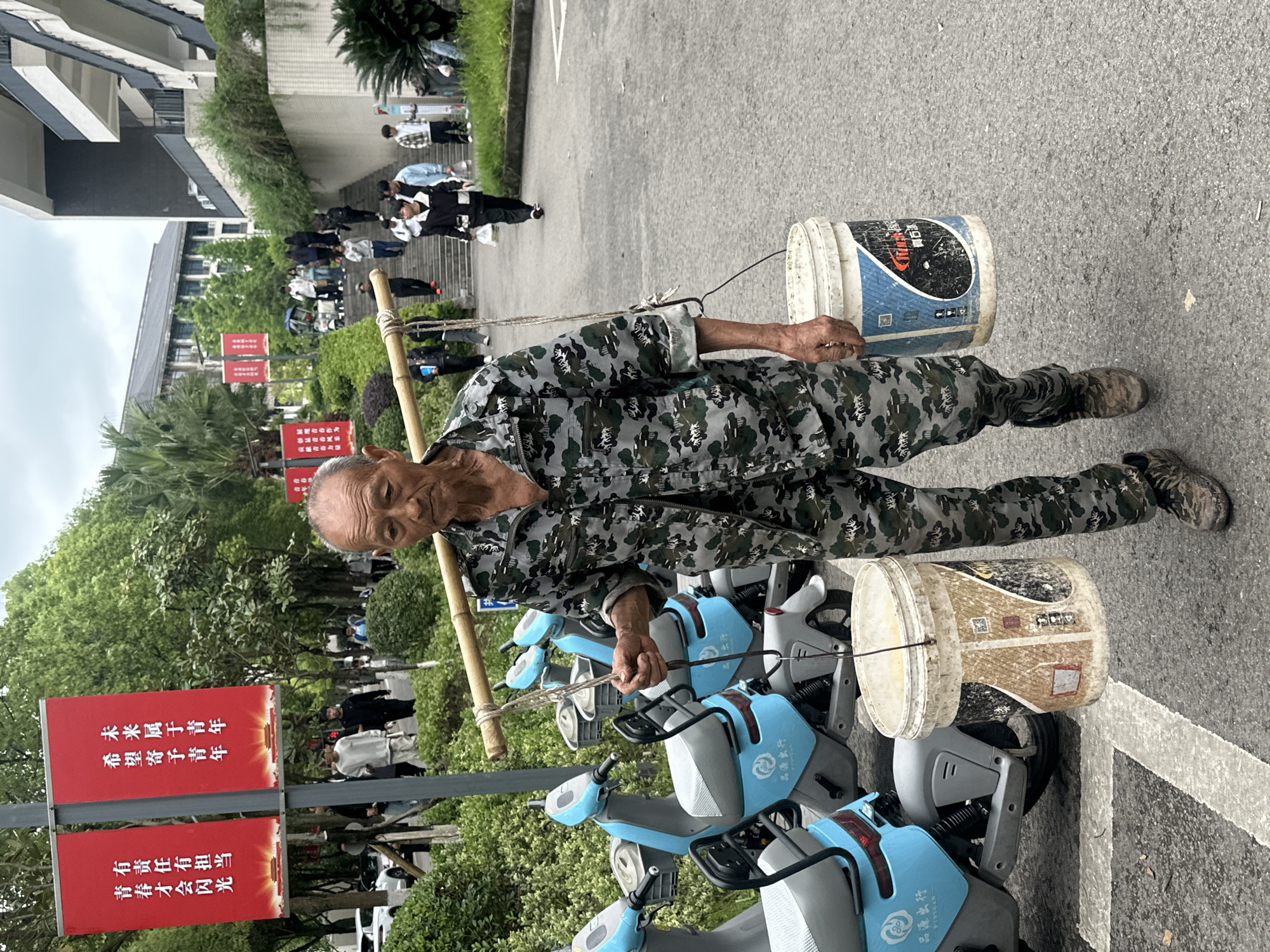 An elderly man is carrying two buckets suspended from a bamboo pole across his shoulders. He is wearing camouflage clothing and walking on a paved surface near a row of light-blue electric bikes. In the background, there is a group of people on stairs leading to a building. Red banners with Chinese characters are on display near the man. This scene is located in Jiangjin, China.