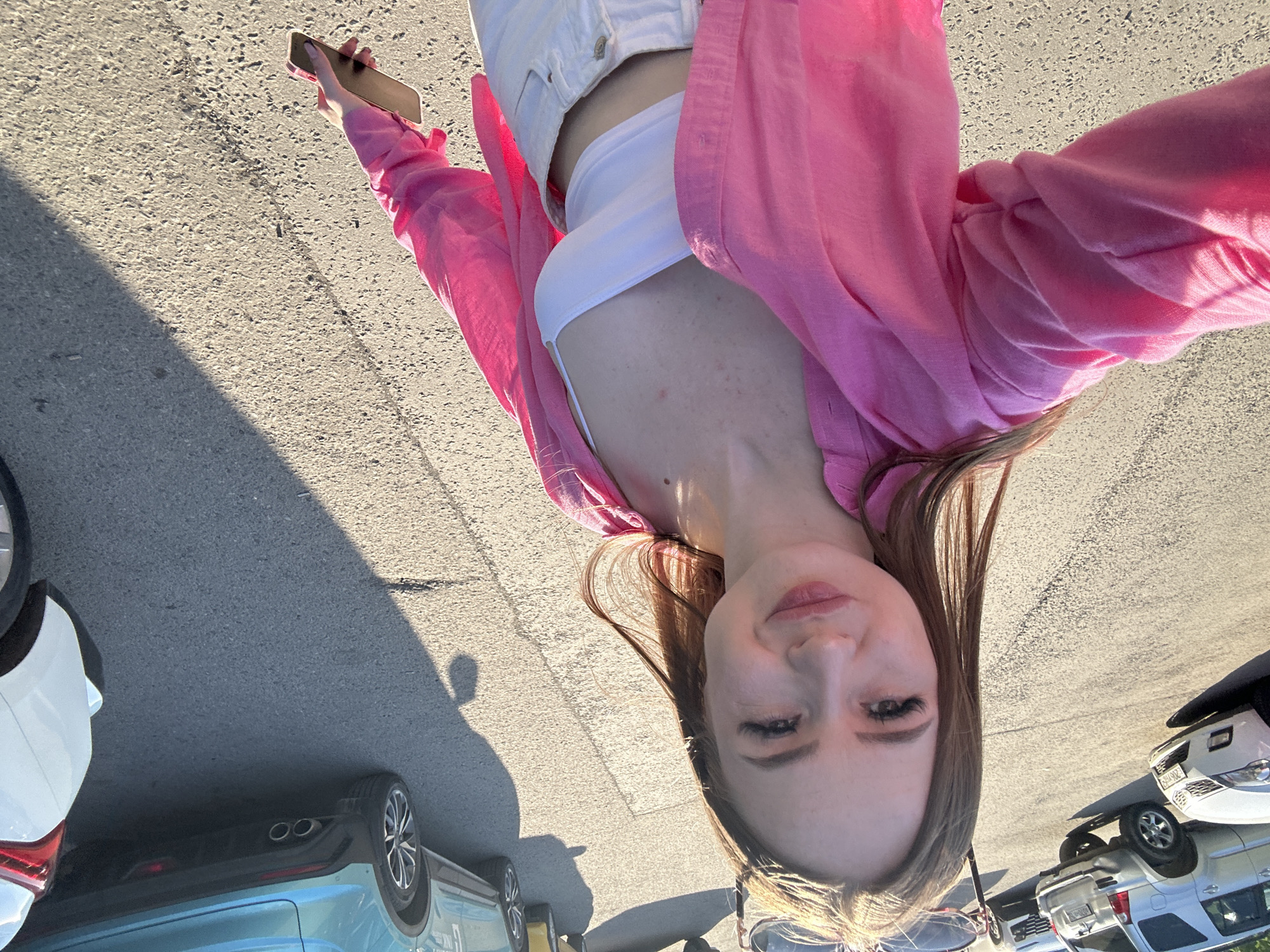 The image displays a young woman taking a high-angle selfie in an outdoor setting, likely a parking lot or paved area. She has long, straight, light brown hair that cascades over her shoulders and a neutral, calm expression as she looks into the camera. She is dressed in a white crop top, layered with a bright pink long-sleeved shirt or light jacket, and white pants or shorts. A dark-colored smartphone is held in her hand, with light-colored manicured nails visible.
The background features an asphalt or concrete surface with various parked cars. A light blue car is partially visible on the left side, and sections of white and silver SUVs are seen on the right. The scene is illuminated by strong, warm sunlight, suggesting either early morning or late afternoon. This is further indicated by the long shadows cast across the ground, including a distinct human silhouette on the right side of the frame. No visible text is present in the image.