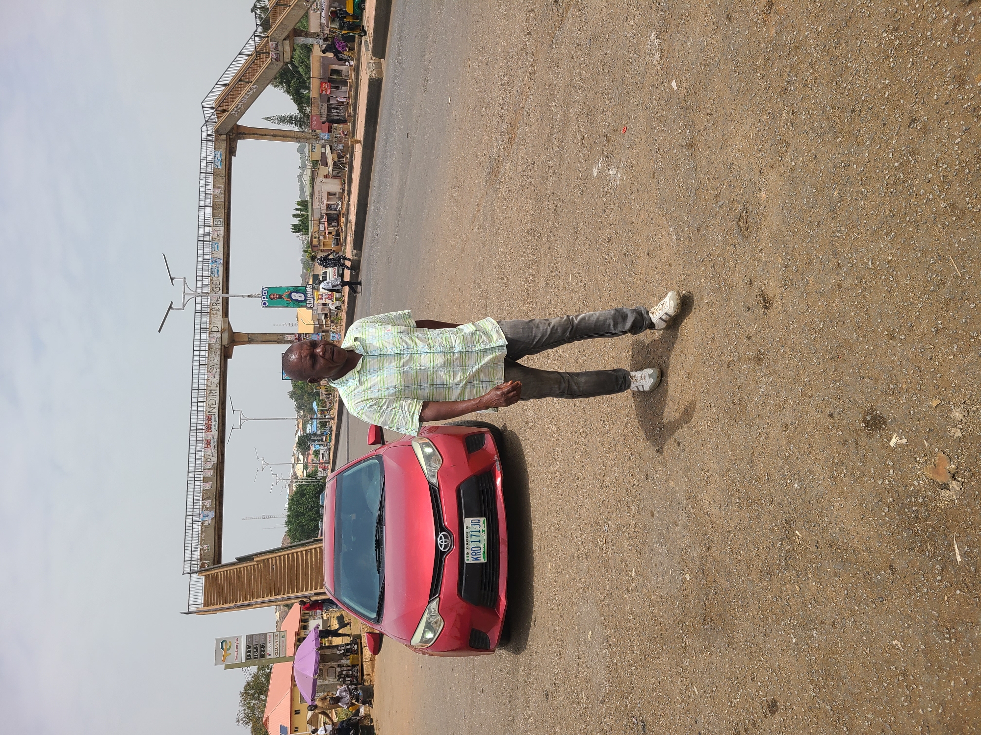 A man stands next to a red Toyota sedan in what appears to be an urban street in Jos, Nigeria. The car has a license plate reading "KRO-171JQ". The man is wearing a light-colored plaid shirt, dark-colored pants, and white sneakers. An elevated pedestrian bridge spans the street in the background, and there are businesses and people visible on both sides of the road. In the lower left, there is a gas station price sign and market stalls with people gathered under umbrellas. The road surface is paved but shows signs of wear and discoloration. The sky is overcast.