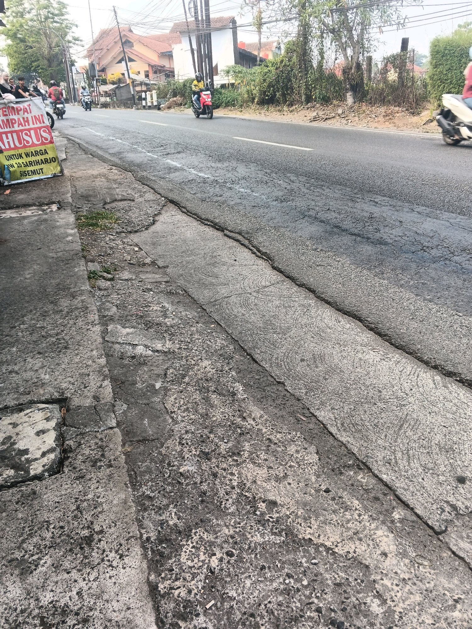 The image depicts a street scene in Ungaran, Indonesia. In the foreground, a cracked concrete sidewalk runs alongside a paved road. The road surface is worn and shows signs of damage. Several motorcycles are visible on the road. One motorcyclist in a yellow jacket is riding a red motorcycle. Another motorcycle, a white scooter, is being ridden on the right side of the road, with its rider wearing a red shirt. In the background, buildings and trees line the street. A sign with Indonesian text is partially visible on the left side of the image. The sky is overcast, suggesting it may be morning or late afternoon. The overall impression is of a busy, everyday street in a bustling Indonesian city.
