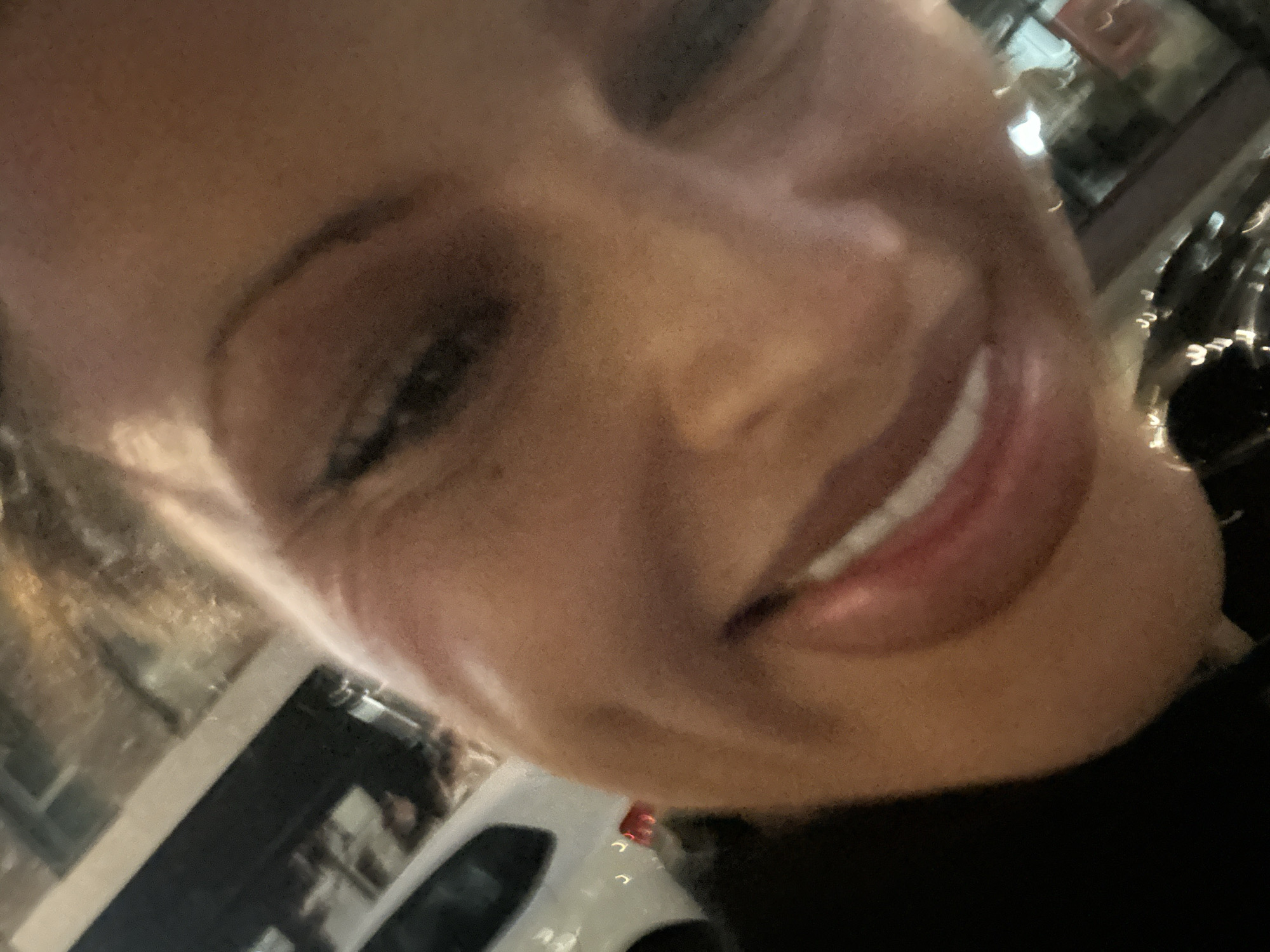 A close-up selfie shows a person's face. They are smiling with their teeth showing. In the background, a vehicle and building are visible, suggesting an urban setting. Based on the presence of a white van and possible architectural details, the location may be within the city of London, United Kingdom.