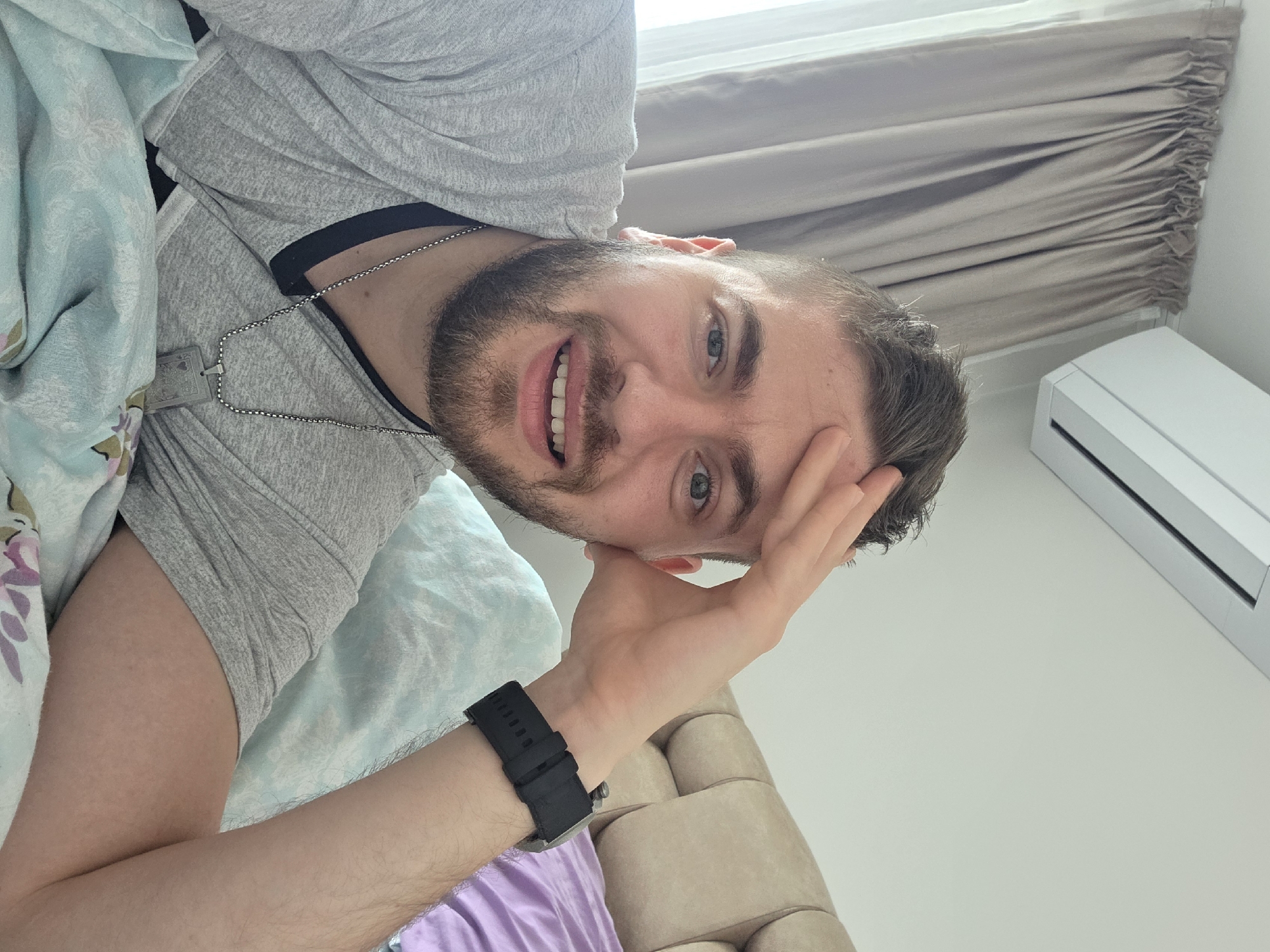 A man lies on a bed with light blue and lilac-patterned sheets. He is wearing a gray t-shirt, and a silver chain necklace. His head is resting on his right hand, and he is smiling. He has a dark beard and short, dark hair. He is wearing a black smartwatch on his left wrist. Behind him, there is a beige curtain and a white air conditioning unit on the wall. The location cannot be determined from the image.