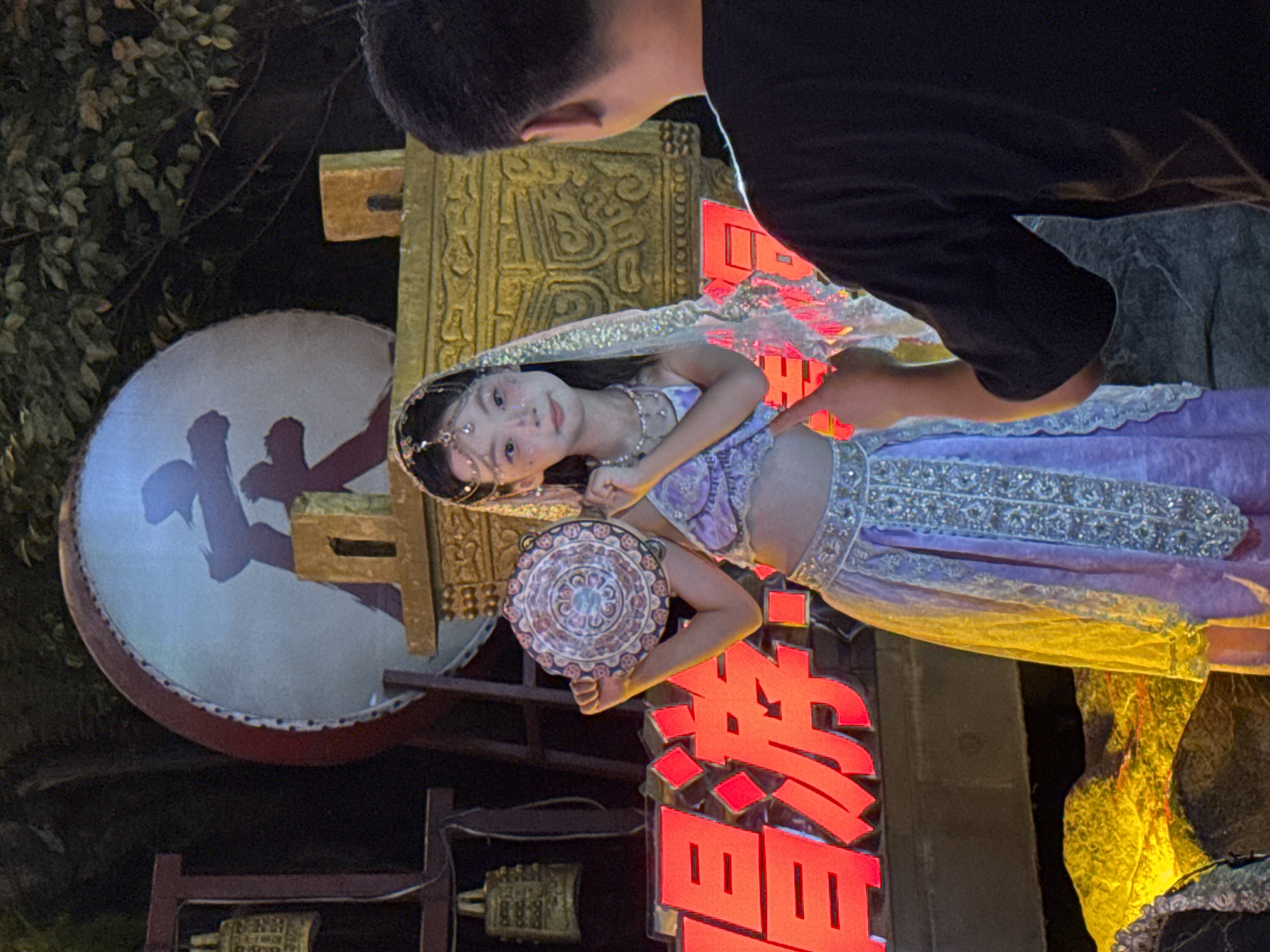 In Yuzhong District, China, a young girl adorned in a traditional purple and gold costume, complete with a beaded headdress and veil, stands on a stage. She holds a circular, decorative tambourine in her right hand. Behind her, a large red drum with Chinese characters is visible, along with an ornate golden structure. In the foreground, a man in a black t-shirt is partially seen from behind, with his right hand extended as if interacting with or directing the girl. The background suggests an outdoor night setting with foliage and illuminated signage displaying large red Chinese characters.