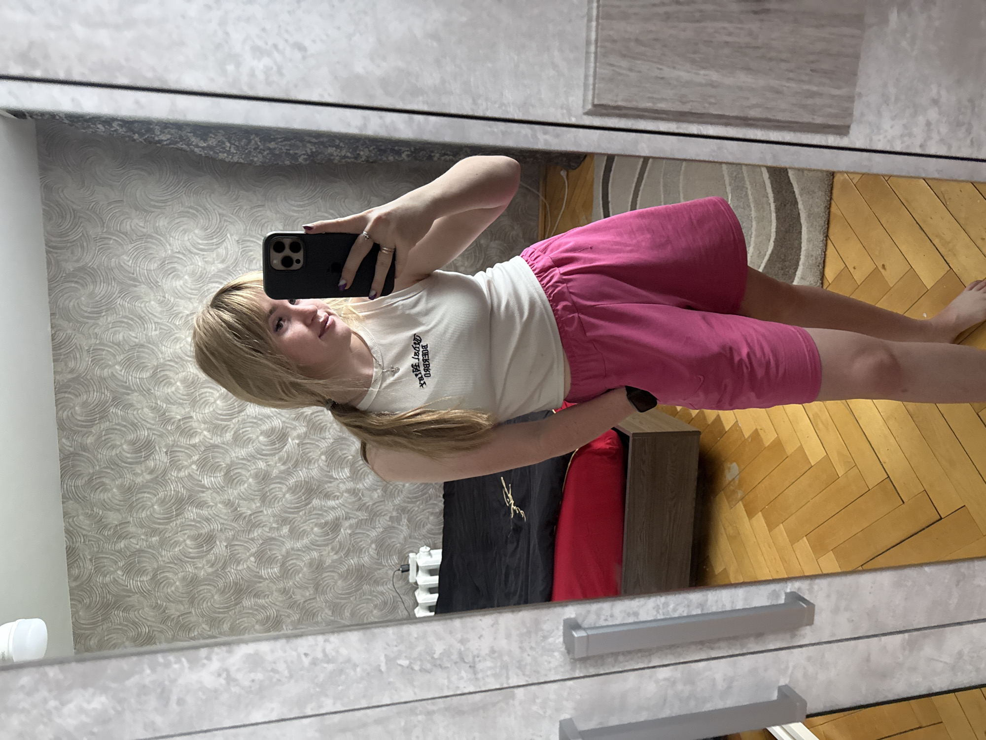 A young woman is taking a selfie in front of a mirror. She has long blonde hair, is wearing a white tank top, and pink shorts. The mirror reflects a room with patterned wallpaper, a wooden floor, and a small cabinet. The location appears to be Марковский, Russia.