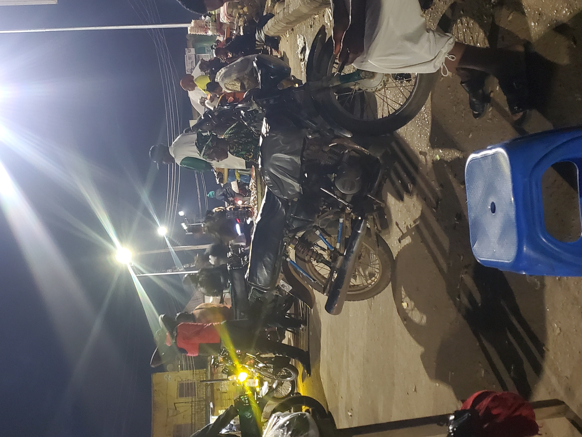 The image depicts a busy street scene at night in Gwong, Nigeria, illuminated by bright streetlights that cast strong shadows. Several motorcycles are parked, some with their headlights on, suggesting a marketplace or gathering area. A variety of people are visible, some standing and talking, others seated.
In the foreground, a blue plastic stool is prominent. To the right, a person wearing white trousers and black sandals is seated, with their legs crossed. A motorcycle is parked behind them. To the left, a red cloth is draped over an unseen object, casting a distinct shadow on the ground.
Further back, more motorcycles are parked, with people gathered around them. One motorcycle in the mid-ground has its headlight on and is being positioned by a person wearing a red shirt and dark trousers. Other individuals, some wearing traditional attire and headscarves, are visible in the background, mingling and interacting. The overall environment appears to be a dusty, outdoor area, possibly a street or a market square, with some buildings and utility poles visible in the distance. The strong lighting creates a dramatic, high-contrast scene.
