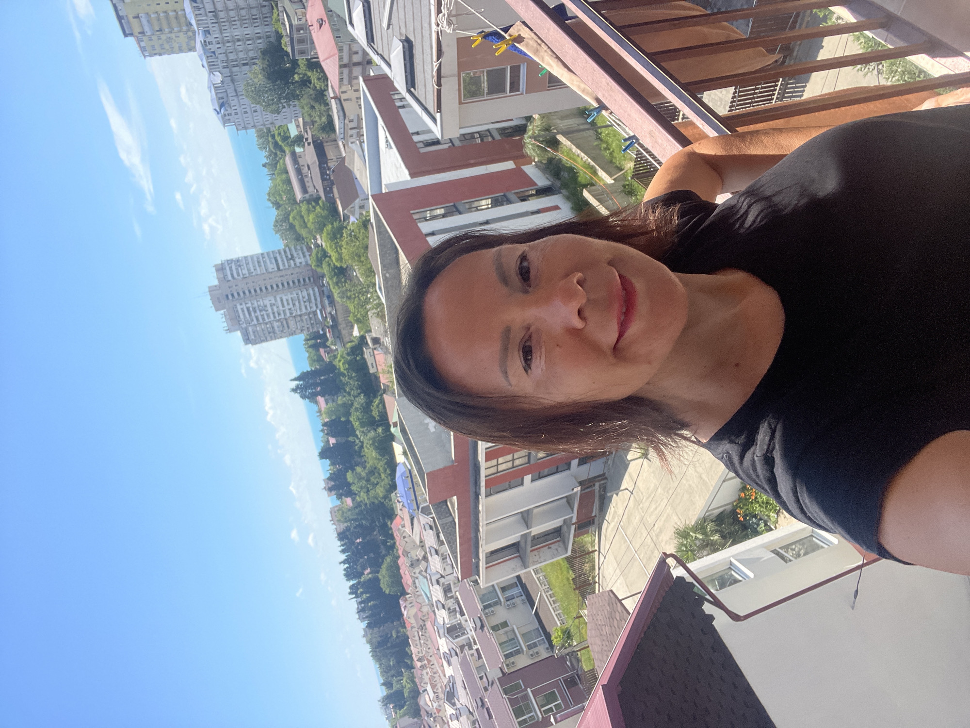 A mid-shot selfie taken from a balcony shows a woman with dark hair, smiling, and wearing a black t-shirt. The background reveals a city skyline with various buildings, including apartment complexes and smaller houses, with the Black Sea visible in the distance. The location appears to be Sochi, Russia, with the characteristic architecture of resort towns. A clothesline with laundry is visible on the right side of the frame, attached to the balcony railing. The sky is clear and blue, indicating a sunny day. The woman's hand rests on her hip.