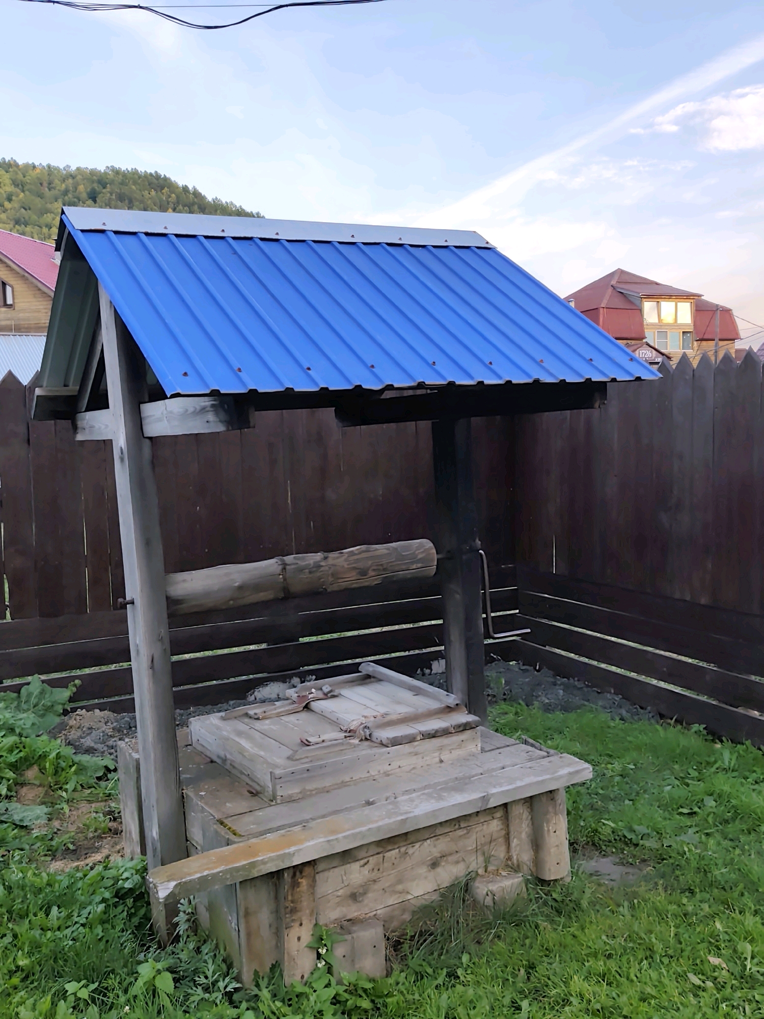 A weathered wooden well with a blue corrugated metal roof stands in a grassy area. The well structure is made of rough-hewn wooden planks, with a larger, aged log acting as a roller for a rope mechanism. A wooden lid covers the top of the well. Behind the well is a dark brown wooden fence. In the background, several houses with red roofs are visible, and a tree-covered hillside can be seen in the distance. The sky is a pale blue with some wispy clouds. The scene appears to be in a rural or semi-rural setting.