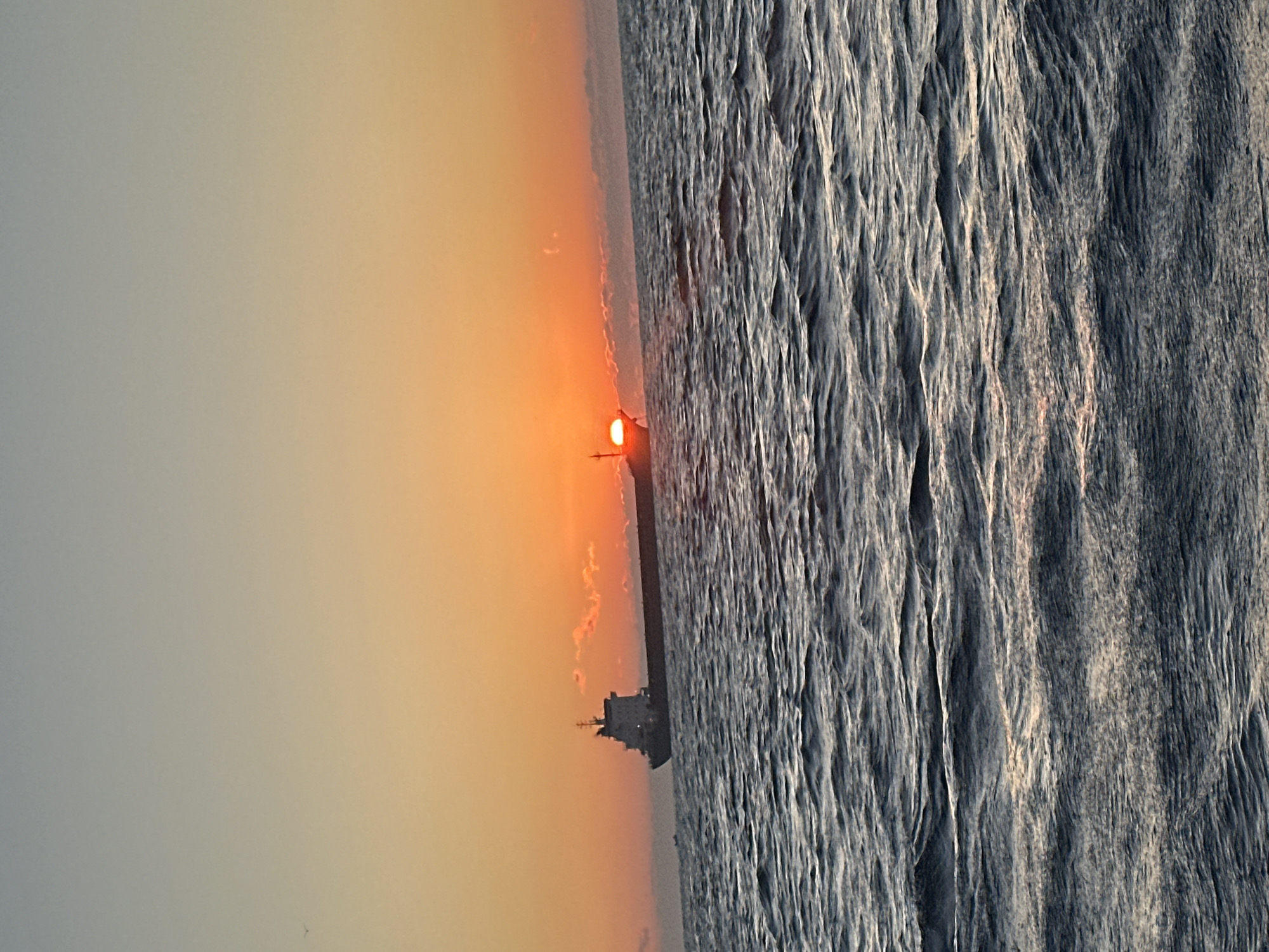 An eye-level, medium shot captures a cargo ship sailing on the ocean at sunset. The sun, a vibrant orange orb, is partially obscured by the horizon, casting a warm glow across the scene. The sky transitions from a soft blue at the top to a fiery orange near the horizon, with hints of pink clouds. The ocean surface is rippled, reflecting the colors of the sky. The cargo ship, dark against the bright sky, is silhouetted, its structure clearly defined. The overall mood is tranquil and serene, evoking a sense of peace and solitude. The location is Unknown, Unknown.