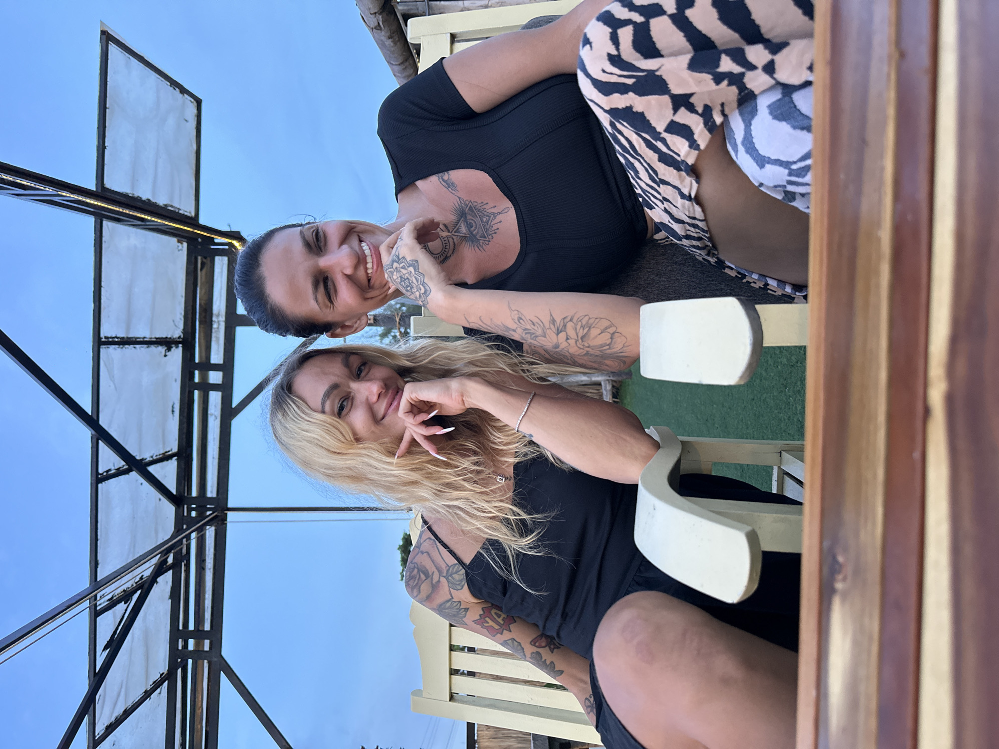 Two women are seated on chairs, possibly at an outdoor restaurant or lounge area, with a table in the foreground and a structure with a canopy overhead. The woman on the left has long blonde hair, is wearing a black dress, and has tattoos on her arms and shoulders. The woman on the right has dark hair, is wearing a black top and a patterned skirt, and also has tattoos visible on her arms and chest. Both women are smiling and looking towards the camera. The background shows a clear blue sky and some greenery.