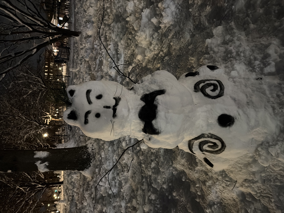 The image captures a whimsical snowman at night, illuminated by ambient light. The snowman is built in three sections, with a distinct character-like appearance rather than a traditional round shape. Its head, the top section, is shaped somewhat like an animal, possibly a cat or bear, with small protrusions resembling ears at the top. The facial features are drawn in black, including two oval eyes, a small triangular nose, a slightly curved mouth, and thin horizontal lines on either side of the face suggesting whiskers.
The middle section of the snowman's body features a large, irregular black patch on its front. The bottom section is adorned with artistic black markings: two prominent spiral or swirl designs and one smaller black dot. Thin, dark branches are used as arms, extending outwards from the middle section of the snowman.
The ground around the snowman is heavily covered in lumpy, undisturbed snow, indicating a recent or significant snowfall. In the background, bare tree branches, some lightly dusted with snow, are silhouetted against the dark sky. Blurred lights, likely from buildings or streetlamps, can be seen in the distance, providing a soft glow that lights the scene. The overall impression is one of a quiet, cold winter night. No discernible text is visible in the image.