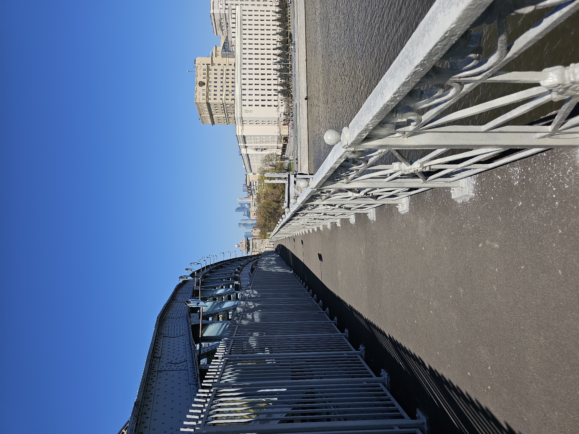 The image depicts a view from a bridge in Moscow, Russia. The focus is on the bridge's infrastructure, including the railings and pedestrian walkway. The architectural elements of the city can be seen in the background, including a large building with numerous windows, likely an administrative or residential structure. The water of the river is visible alongside the bridge. The sky is clear and blue. The scene suggests a sunny day in an urban setting.