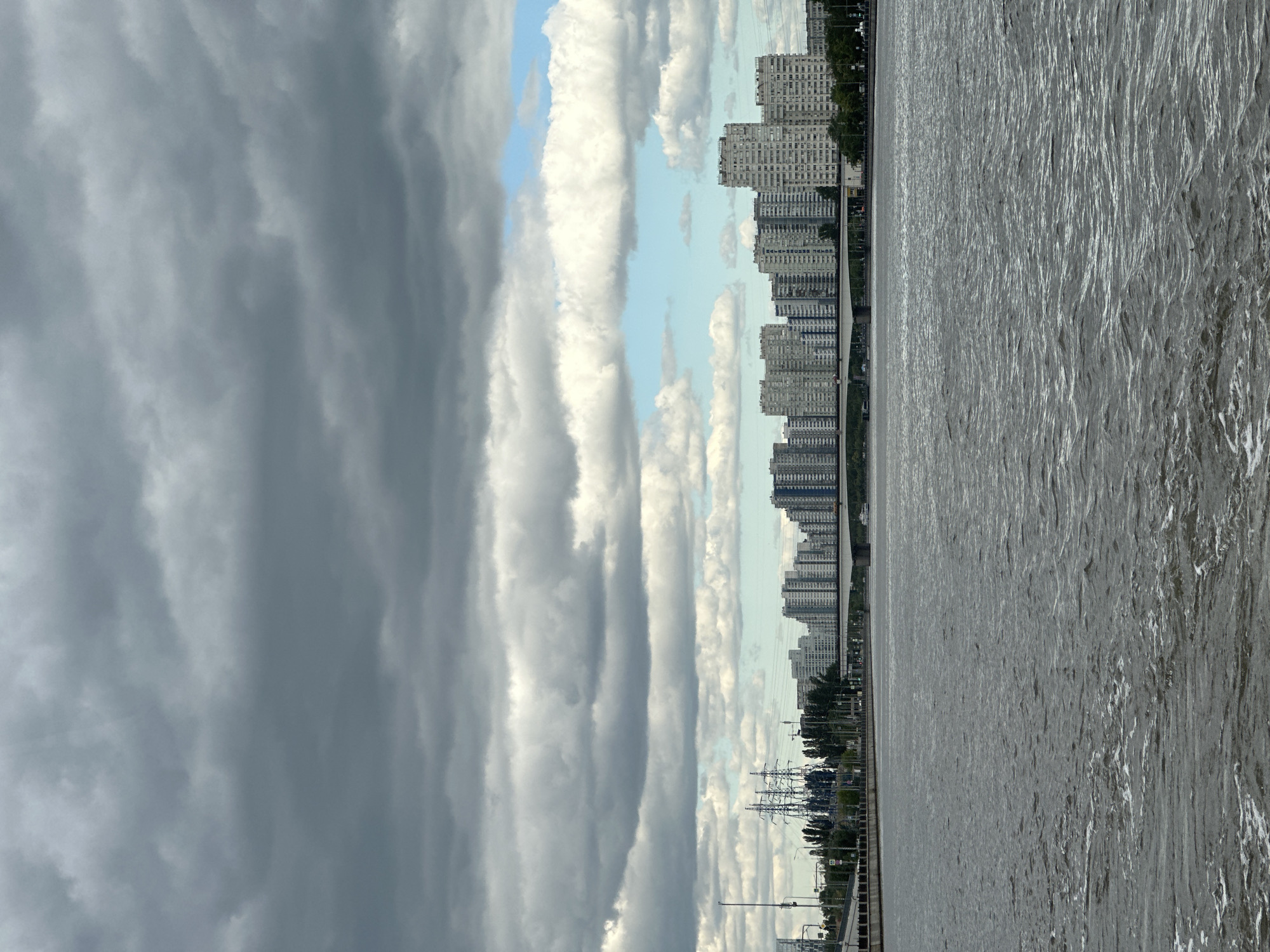 The image shows a wide river in Moscow, Russia, with a cityscape on the horizon. The sky is overcast with dark gray clouds, but there are patches of blue sky visible. On the riverbank, there are tall apartment buildings, a bridge, and power lines. The water is choppy, with small waves and white foam on the surface, suggesting it may be windy. The overall scene depicts an urban environment on a cloudy day.